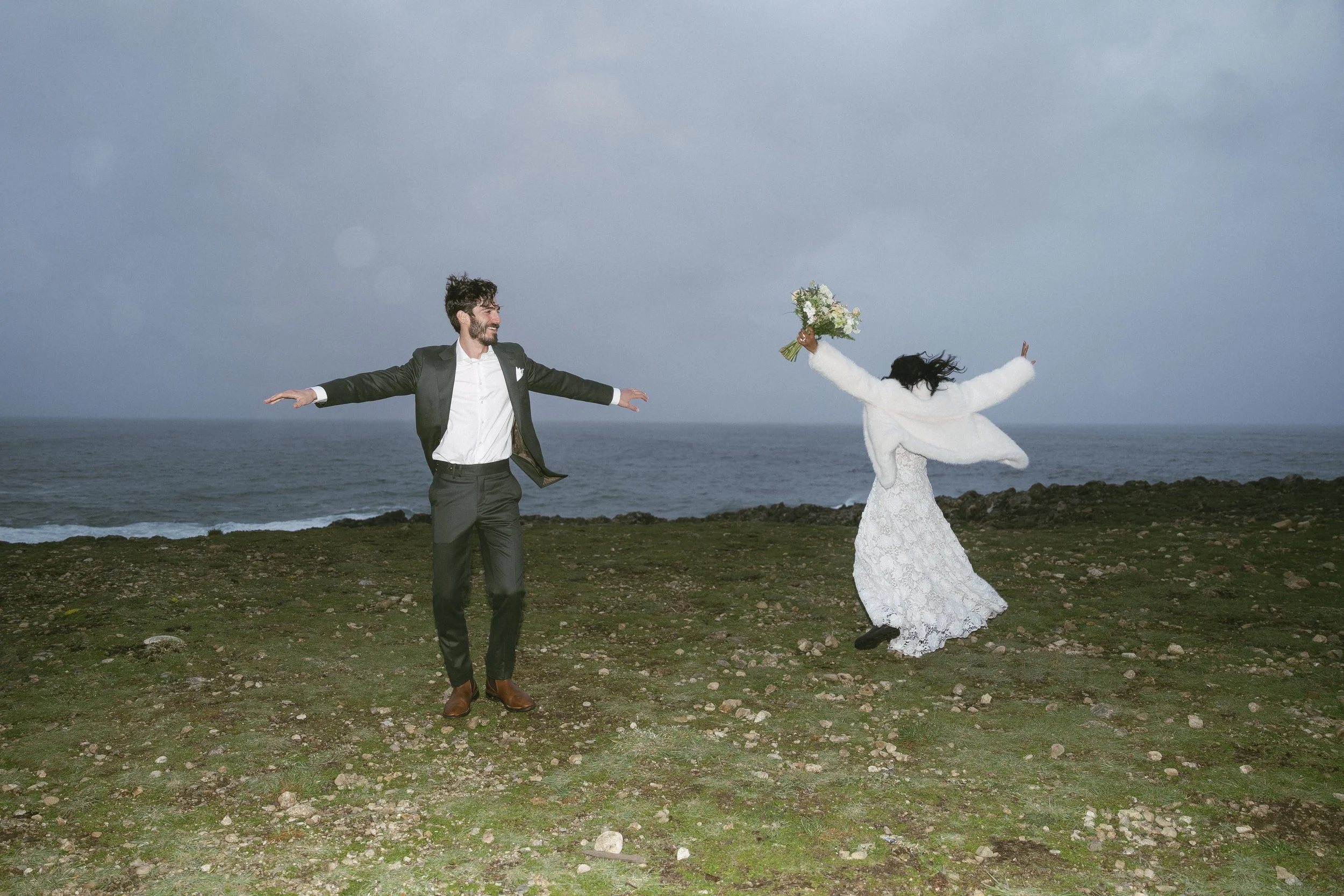 A newlywed couple dancing by the sea on a grassy area with rocks, in formal attire. The groom has his arms outstretched and is smiling, while the bride, holding a bouquet, jumps in the air with her arms raised.