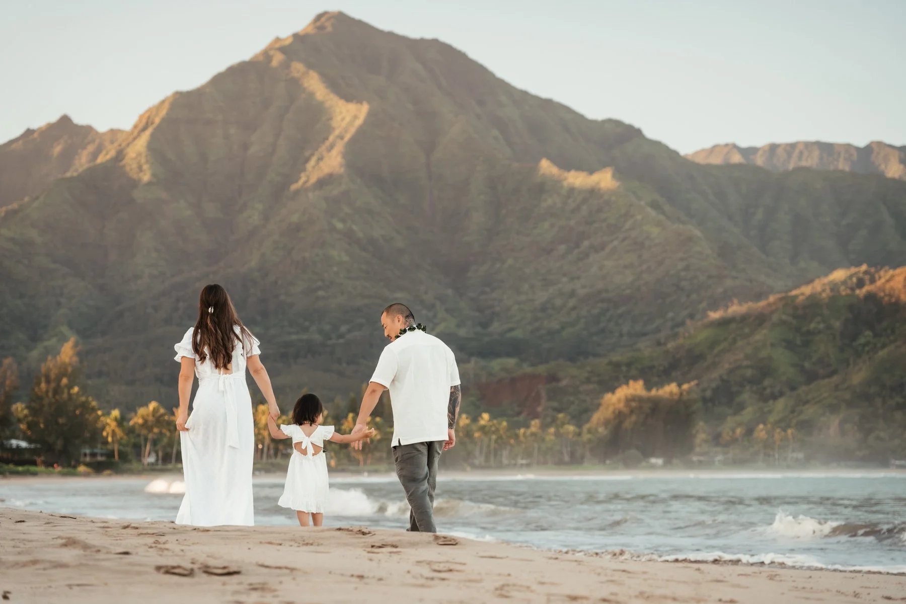 A happy family of three with a man, woman, and girl at the beach during daytime with water and distant mountains in the background.