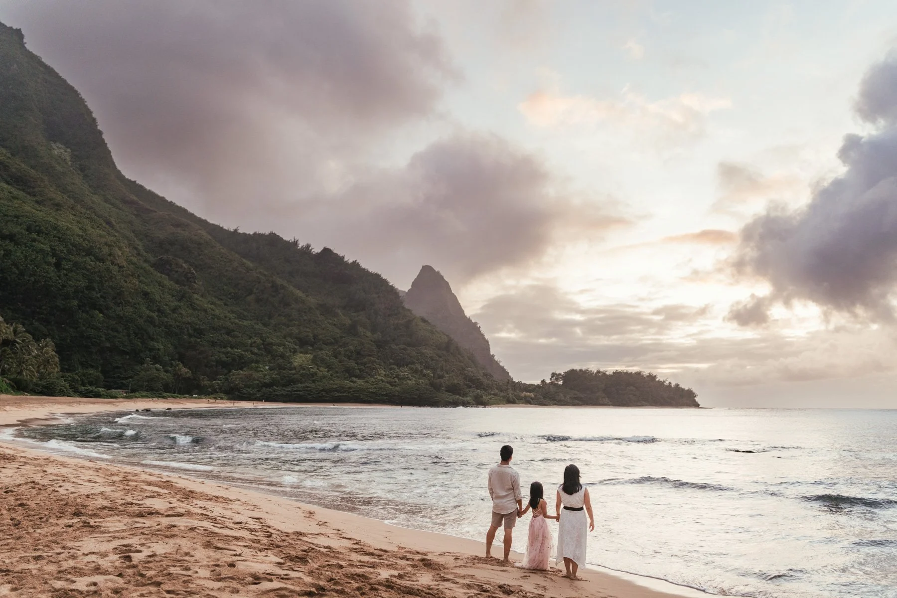 Family of three walking along the shoreline of Haena Beach on Kauai at sunset, with green mountains and soft clouds behind them