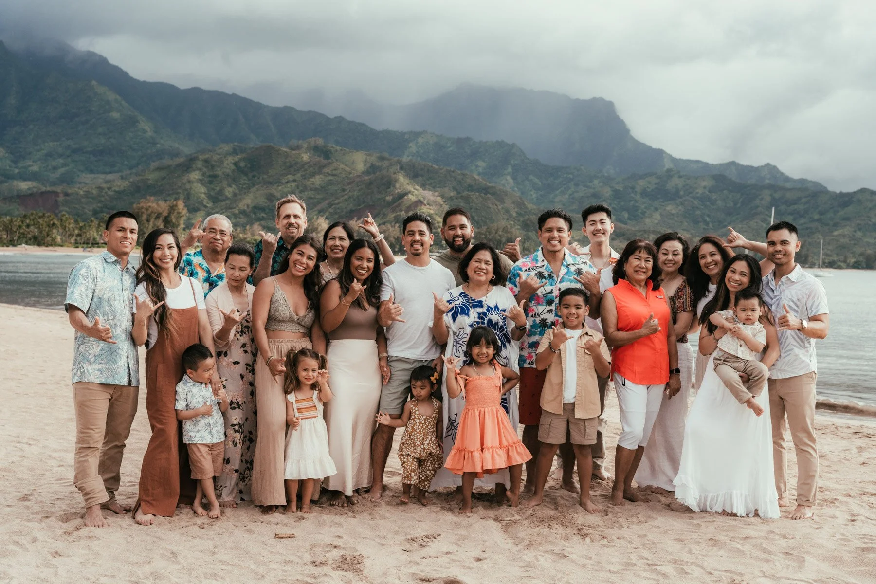 Large multigenerational family of about 30 people posing together on Hanalei Beach on Kauai with green mountains behind them