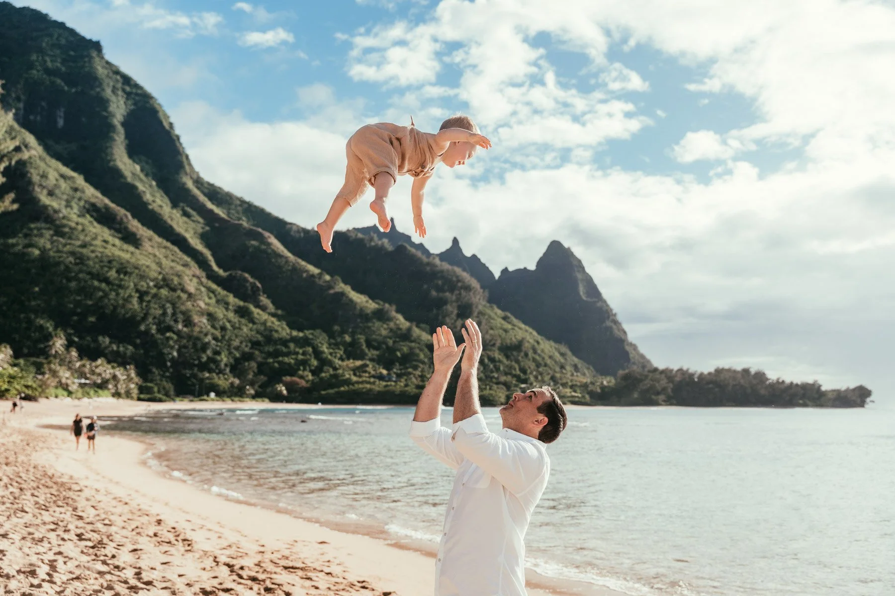 Father tossing a smiling baby into the air on a Haena beach on Kauai, with sharp green mountain ridges and blue sky behind them