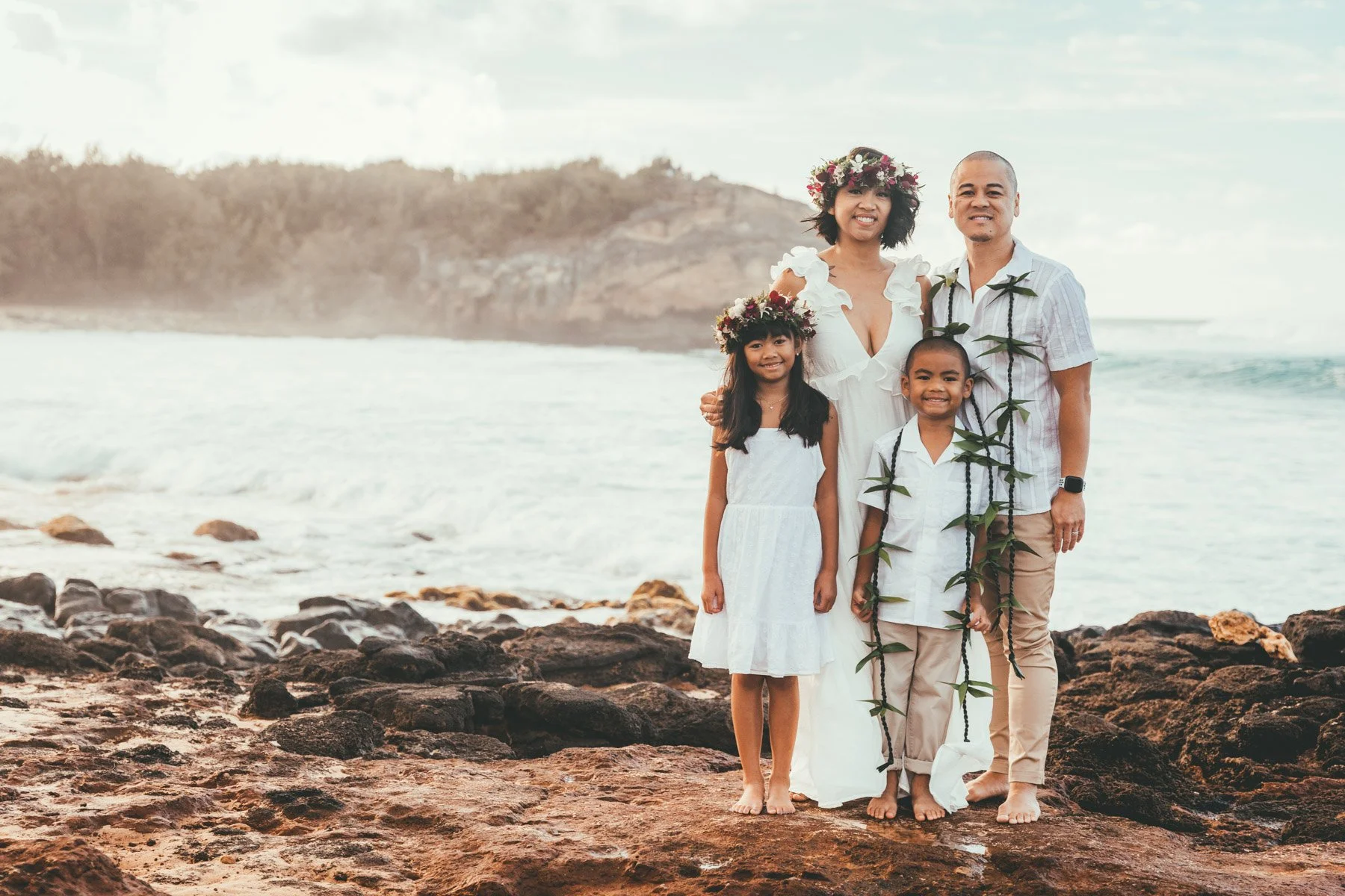 Family of four with a girl in a flower crown and a boy in a leaf lei standing together on lava rocks at Shipwrecks Beach in Poipu on Kauai's South Shore, with ocean waves behind them