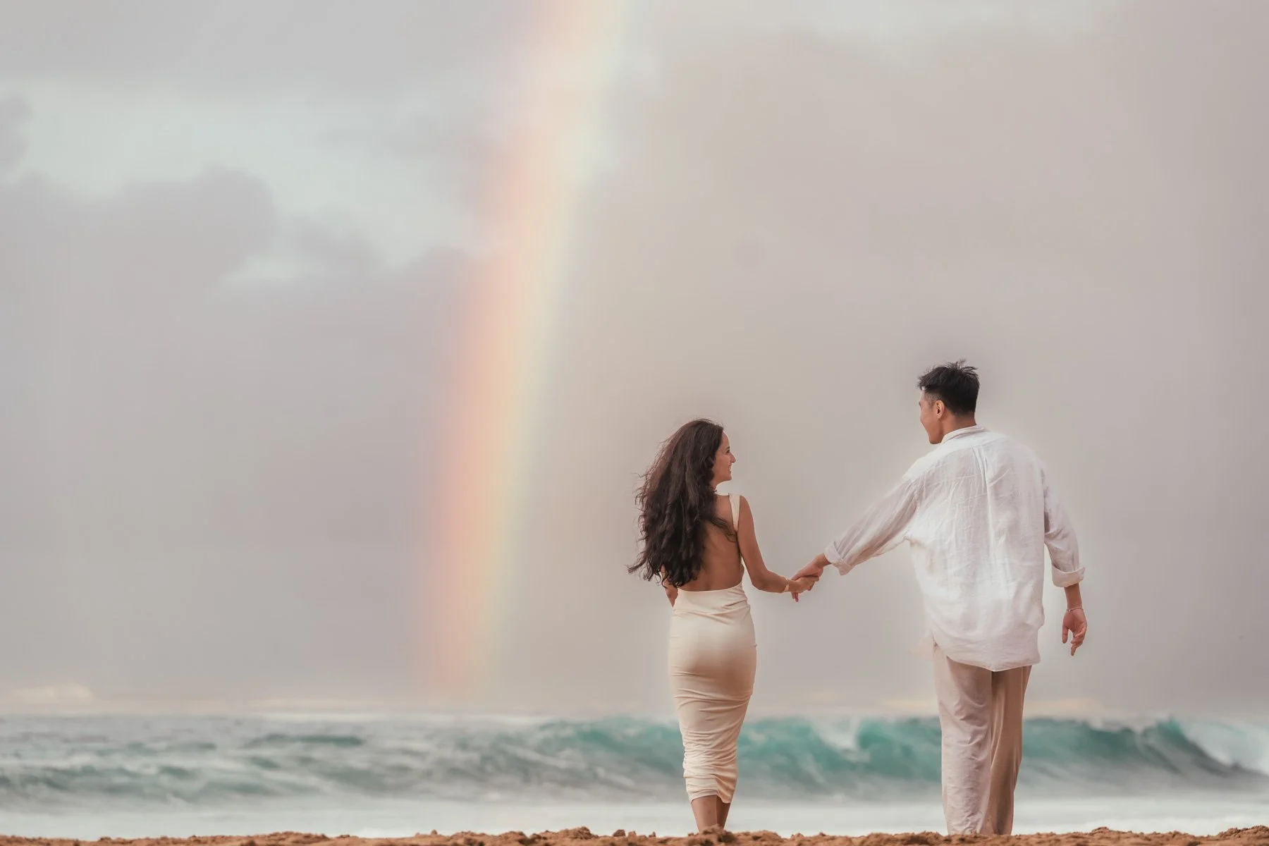 couple walking hand in hand towards rainbow over the ocean