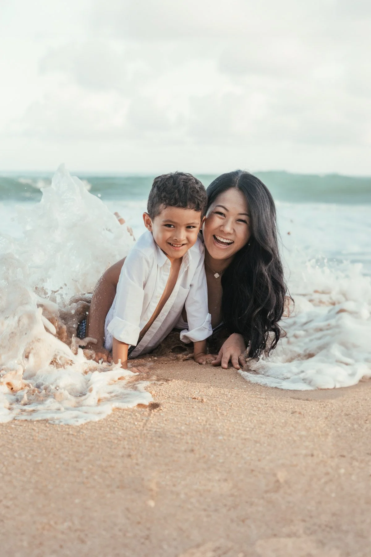 Mother and young son lying in the sand and laughing as a wave washes over them on a Kauai beach during a family photo session