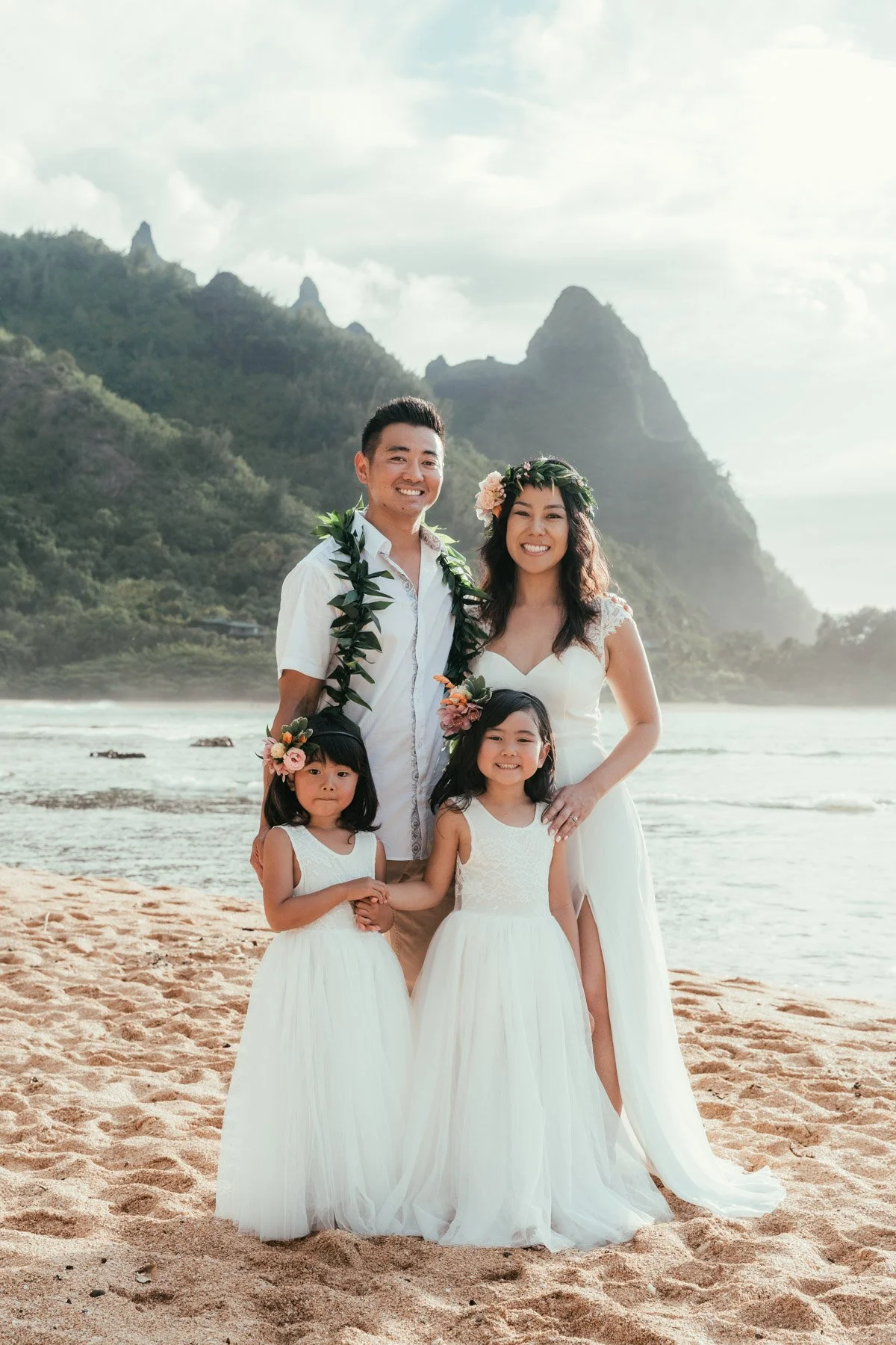 Smiling family of four in white with leis and flower crowns, two young daughters in front, standing on a Kauai beach with green North Shore mountains behind them