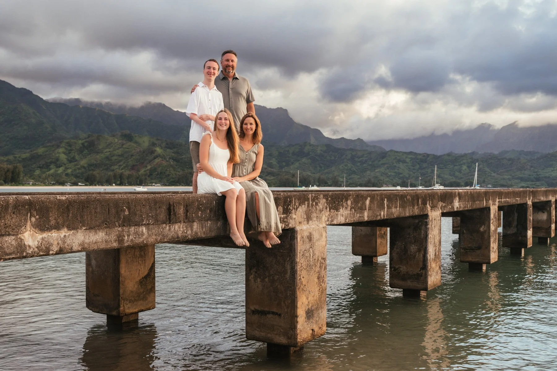 Parents and two children in white outfits posing together on the Hanalei Pier on Kauai's North Shore, with green mountains and cloudy sky behind them