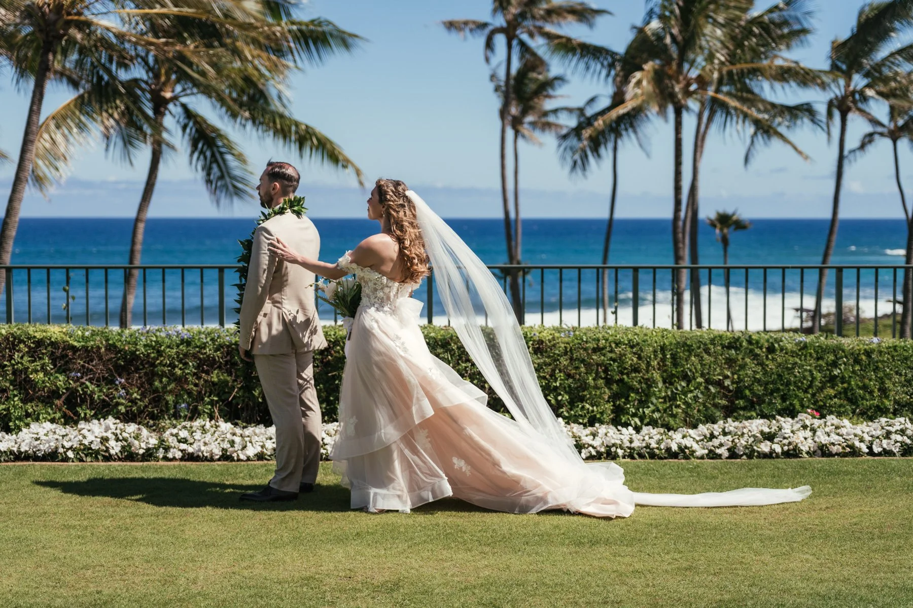 Bride in a flowing veil touching the groom's shoulder during a first look on the oceanfront lawn at the Grand Hyatt Kauai Resort in Poipu