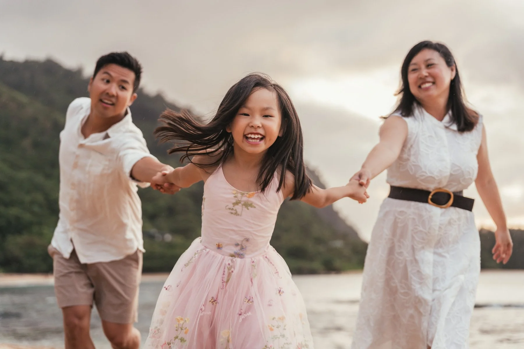 Father, daughter in a pink floral dress and mother holding hands and laughing on a Kauai beach during a family photo session, with green mountains behind them