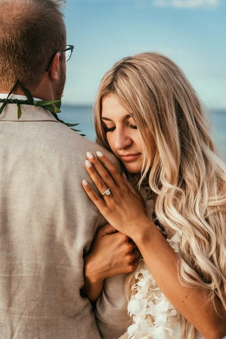 A woman with long blonde hair embraces a man, showing her engagement ring on her left hand, with a body of water in the background.