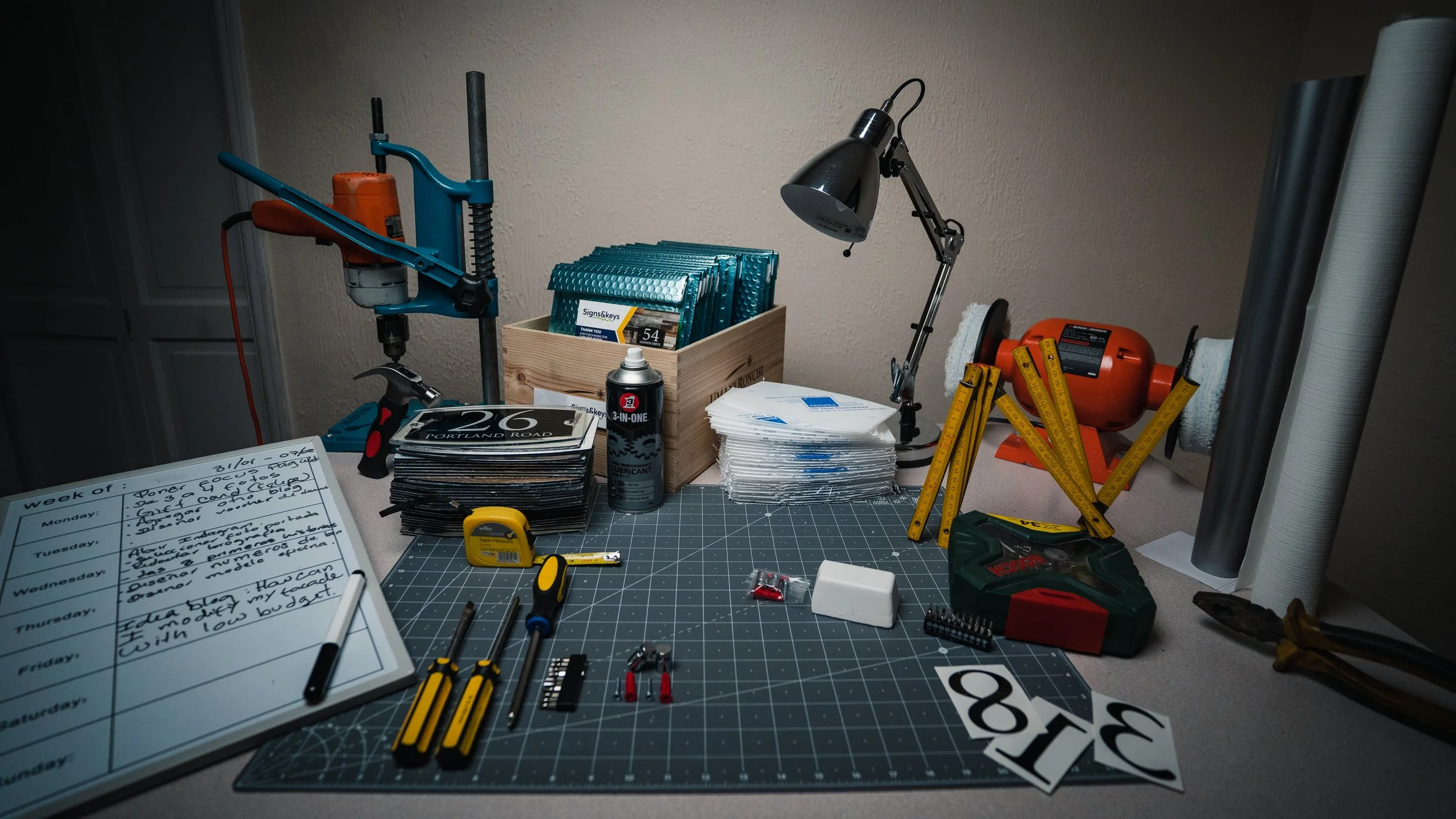 A cluttered workbench with tools, a tape measure, a glue gun, screws, a magazine, a notebook, a pair of pliers, a ruler, a saw blade, a paper cutter, a lamp, a box of files, a stack of papers, a black cutting mat, a bag of screws, and an orange bench grinder.