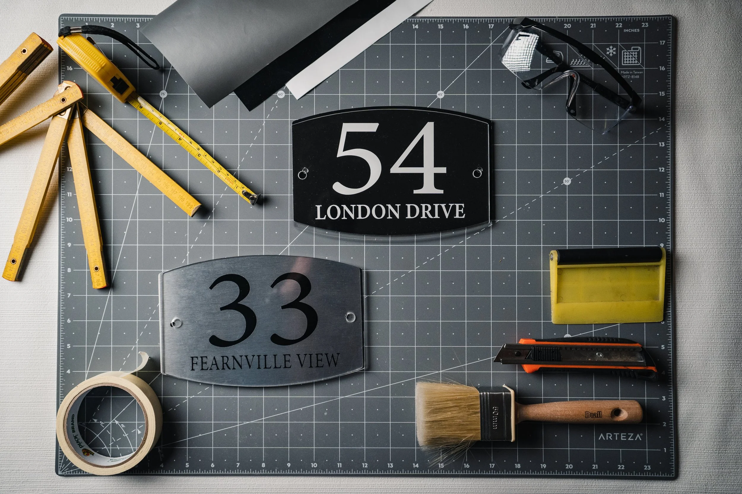 Collection of tools and signs on a work cutting mat, including a paintbrush, a yellow tape measure, a black and yellow utility knife, a yellow level, a pair of goggles, a roll of masking tape, and signs with addresses '54 LONDON DRIVE' and '33 FEARNVILLE VIEW'.