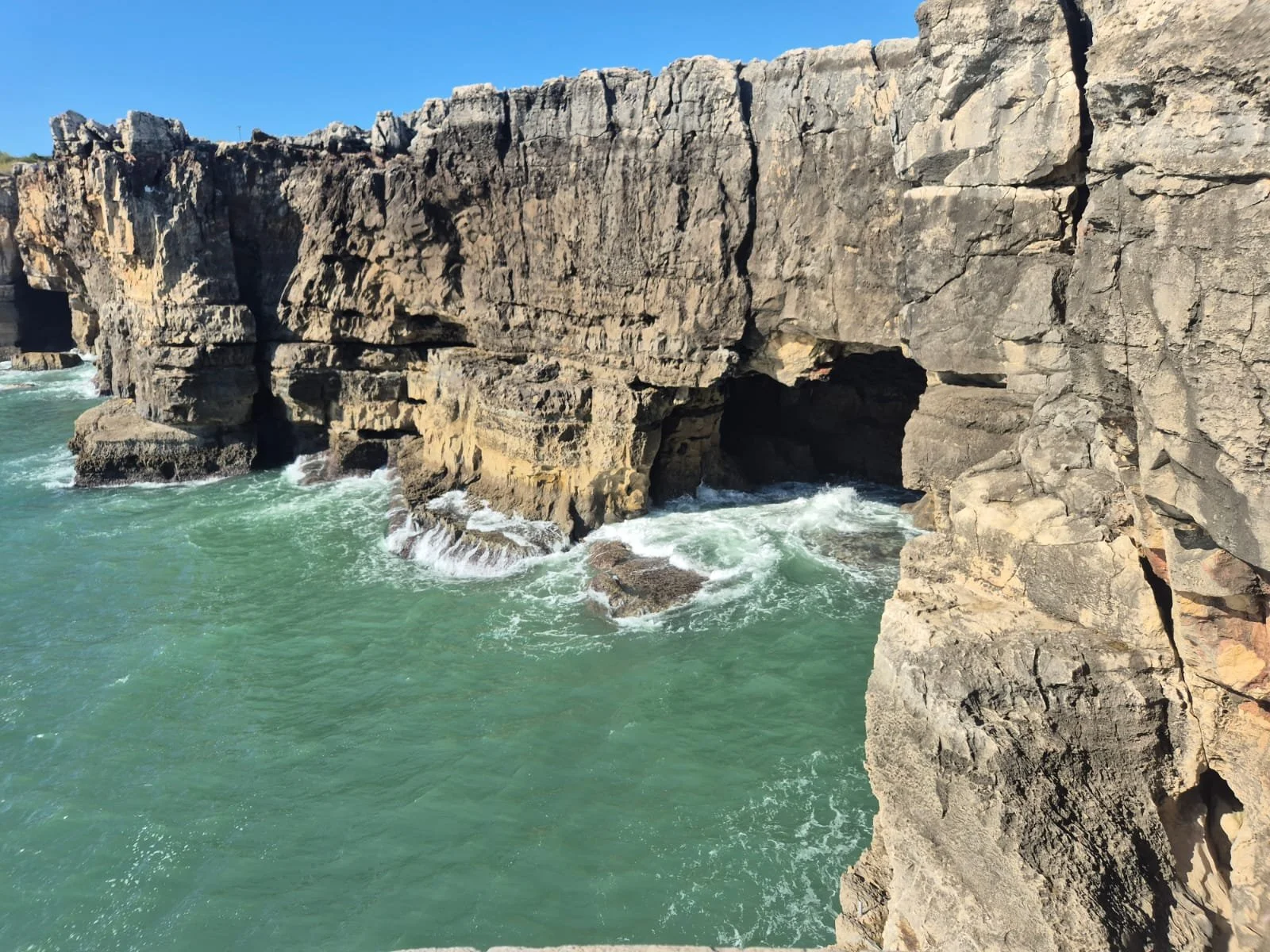 Cliffside with caves and rocks along a turbulent green ocean under a clear blue sky.