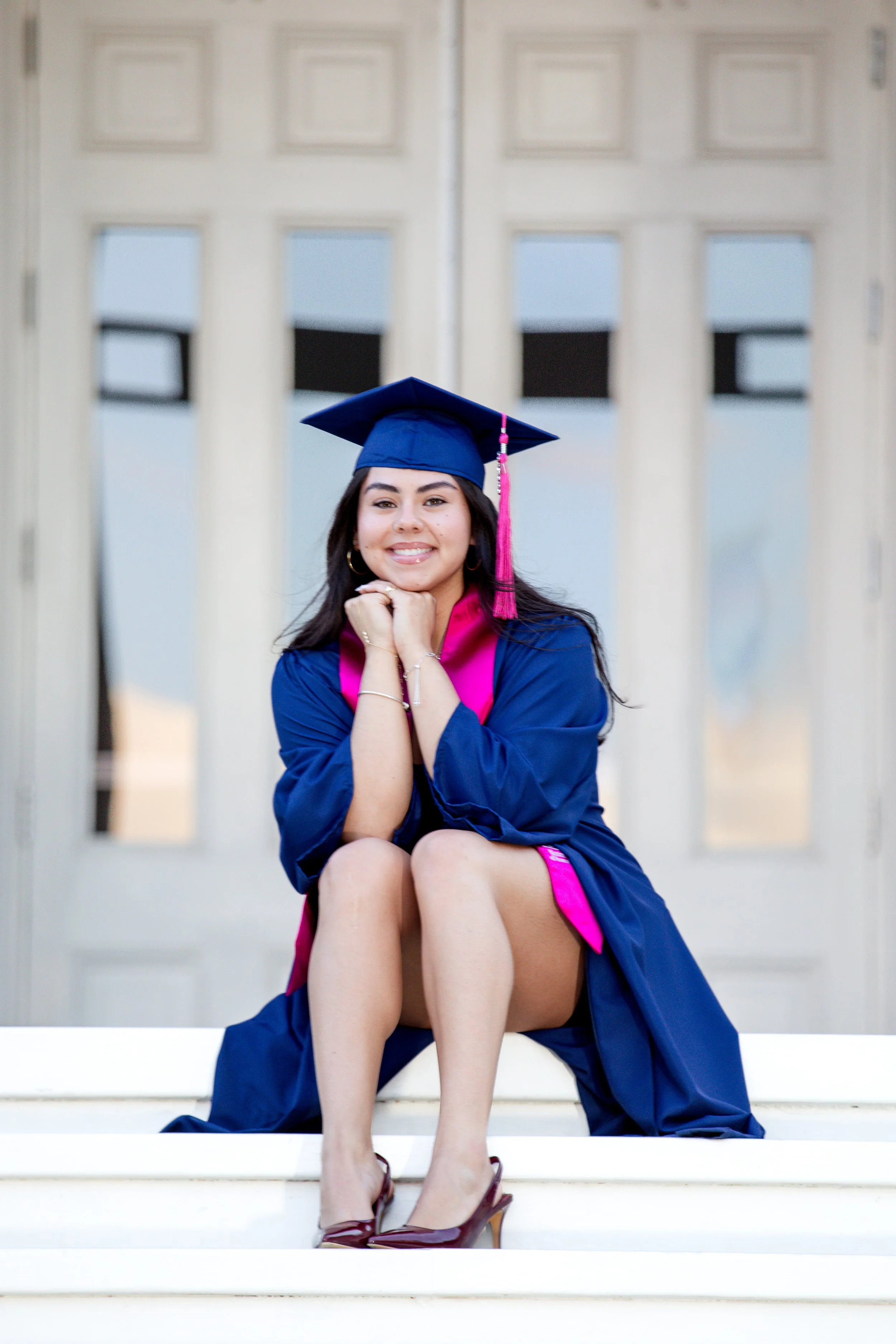 A young woman in a blue graduation gown and cap, with pink accents, sitting on steps with her knees up, smiling with her chin resting on her hands.