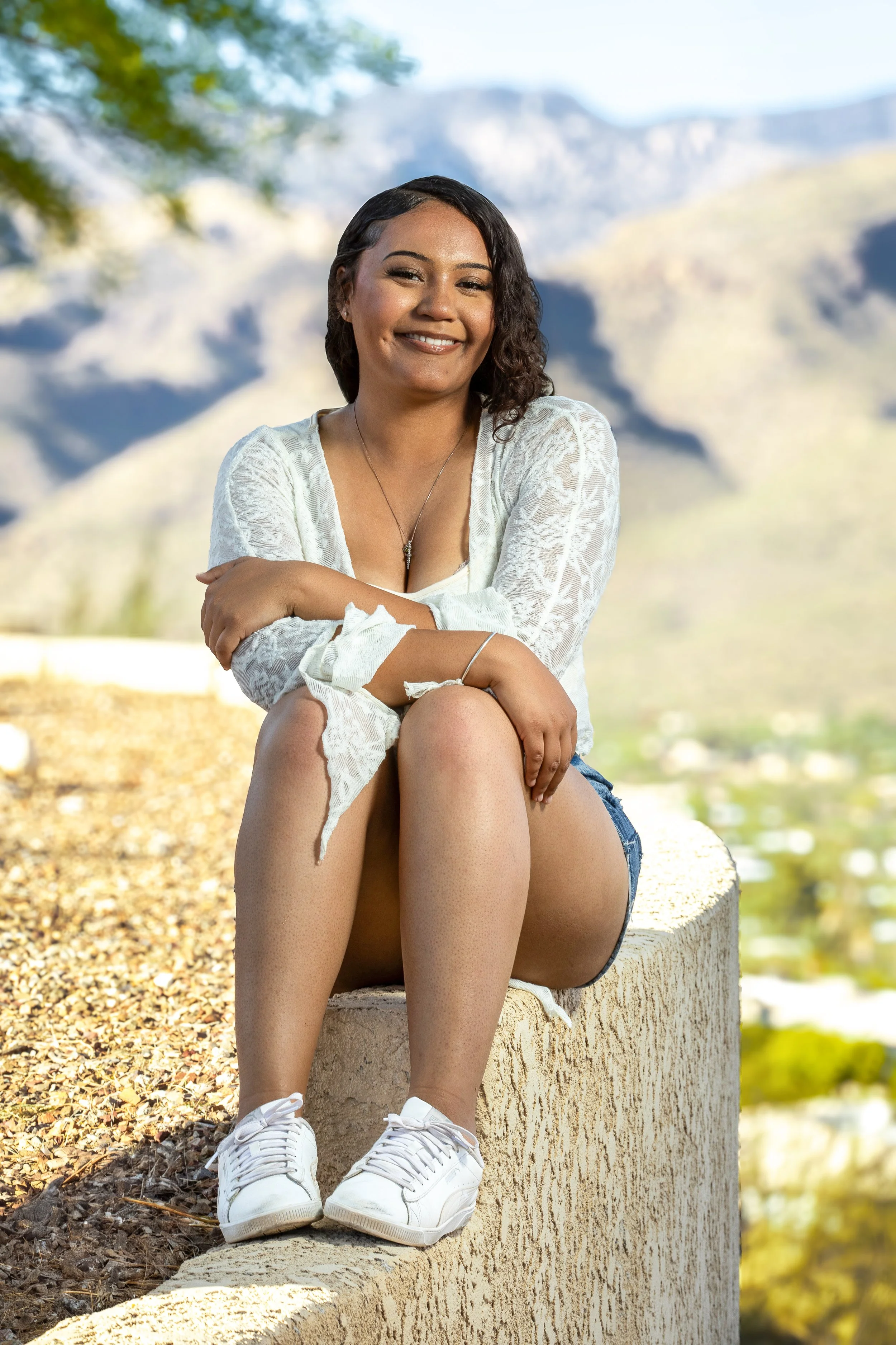 Young woman sitting on a ledge outdoors with mountains and a valley in the background, smiling and looking at the camera.
