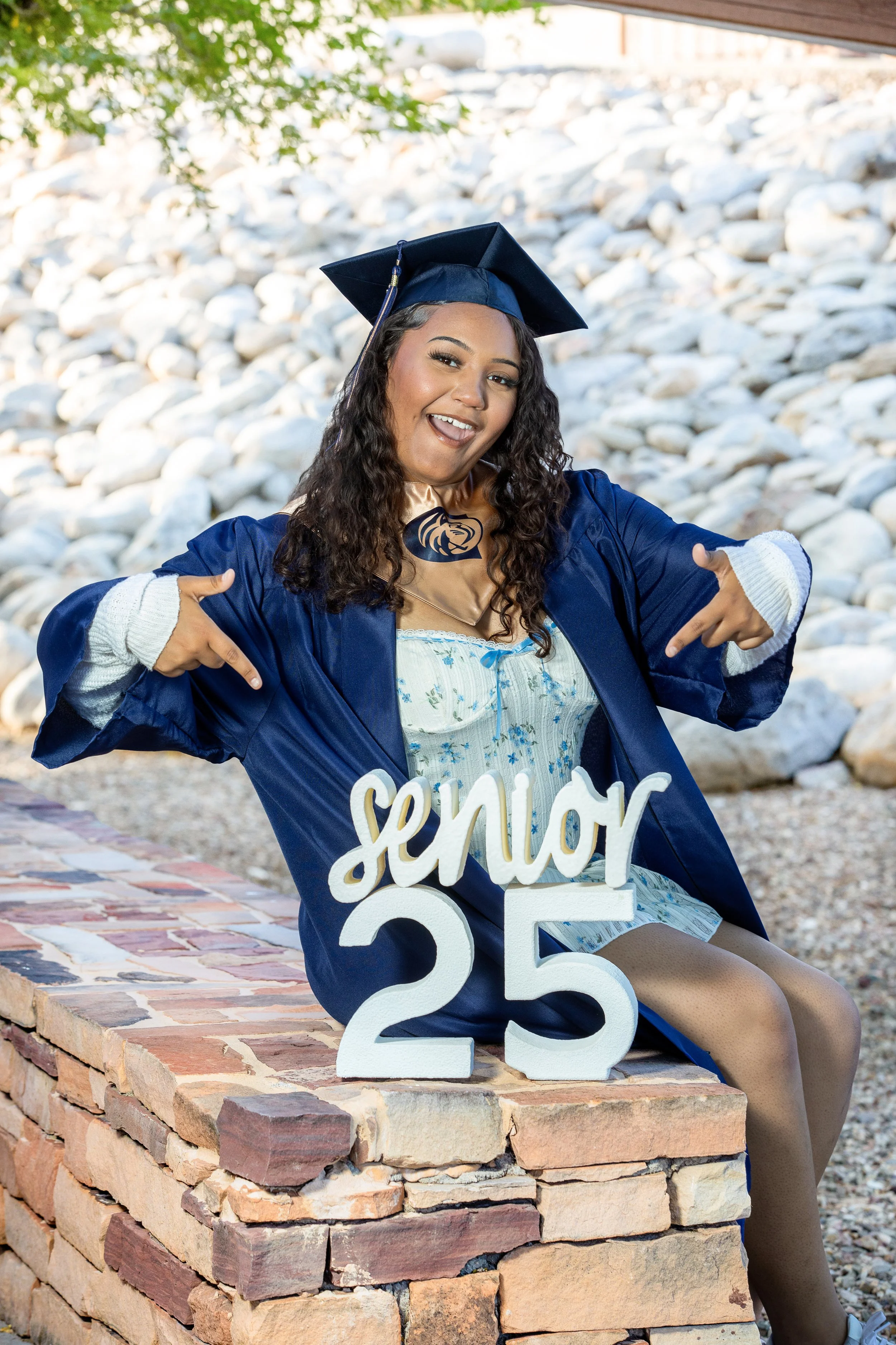 A young woman in a graduation gown and cap, sitting on a brick ledge, pointing to a sign that says 'senior 25' in front of a rocky area.