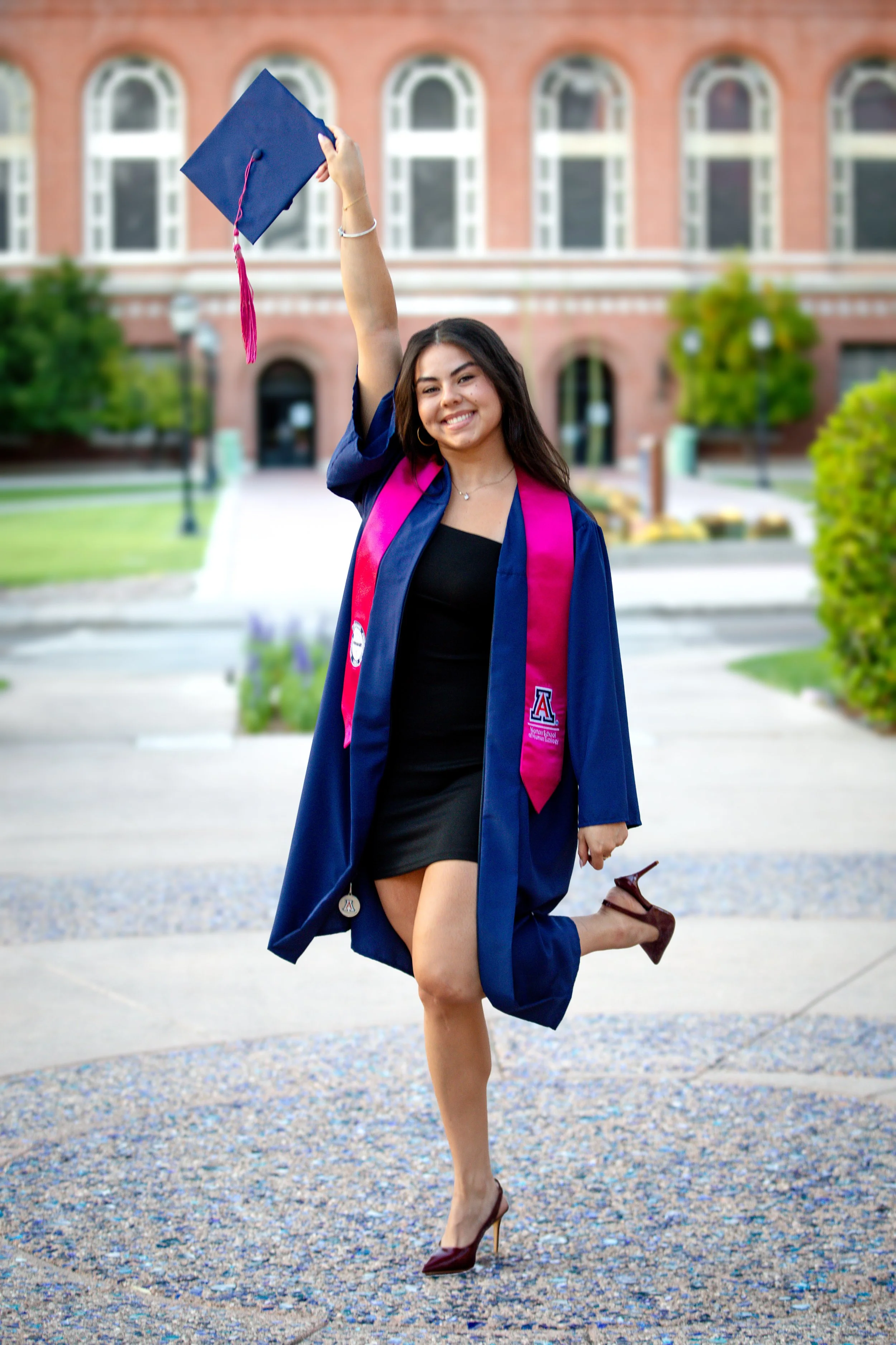 Young woman in a graduation gown and cap holding her cap in the air, standing on a college campus.