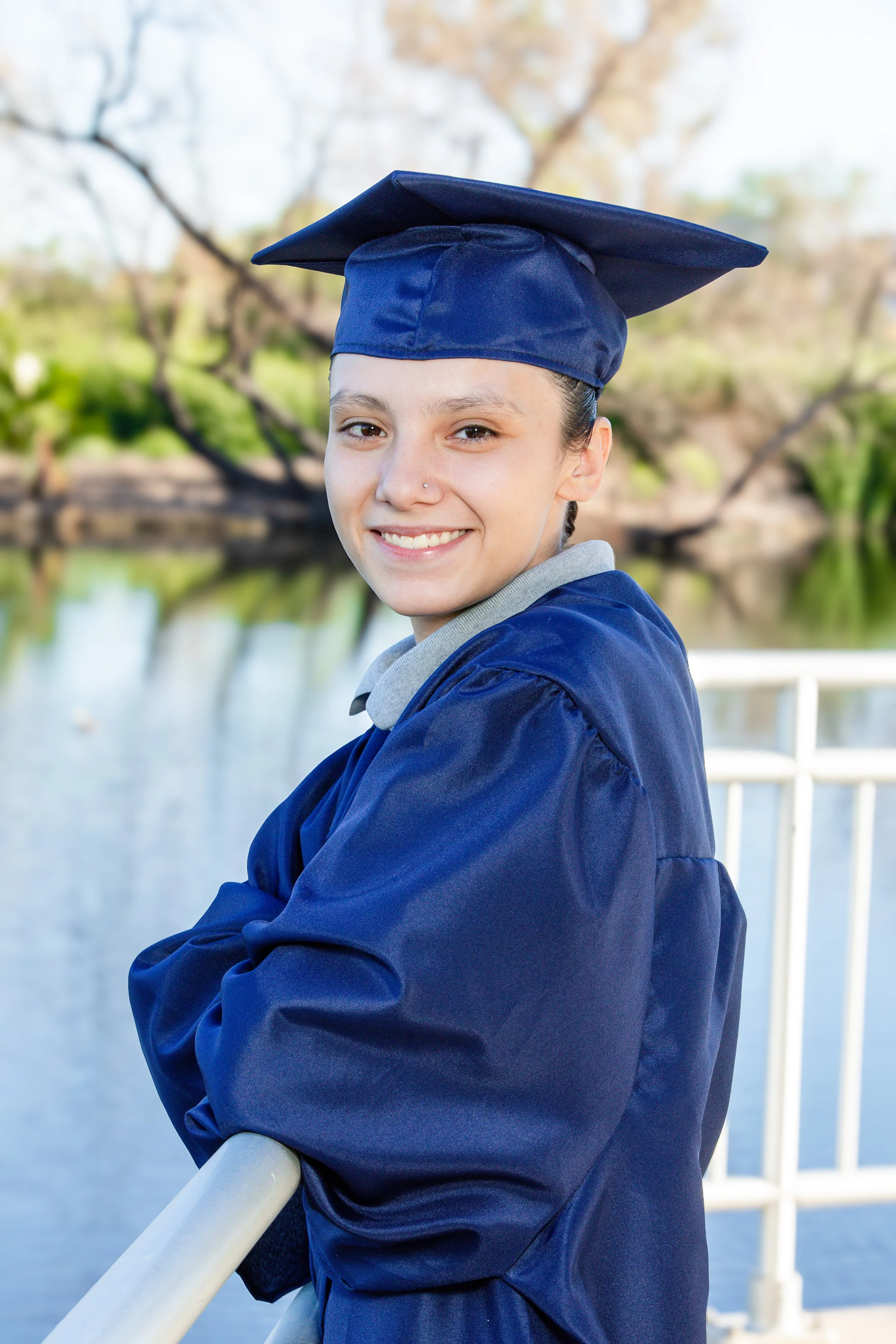 Young woman wearing a graduation cap and gown smiling outdoors near a body of water.