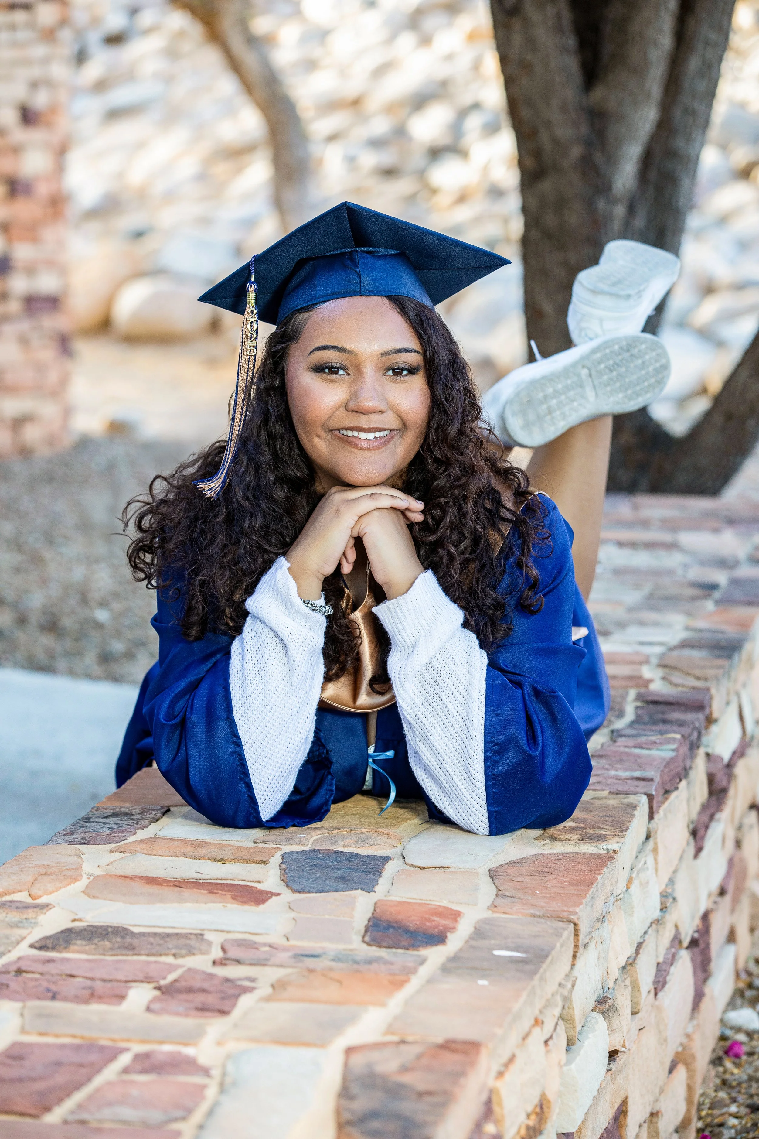 Young woman in a blue graduation cap and gown, smiling and resting her chin on her hands, lying on a brick wall outdoors.