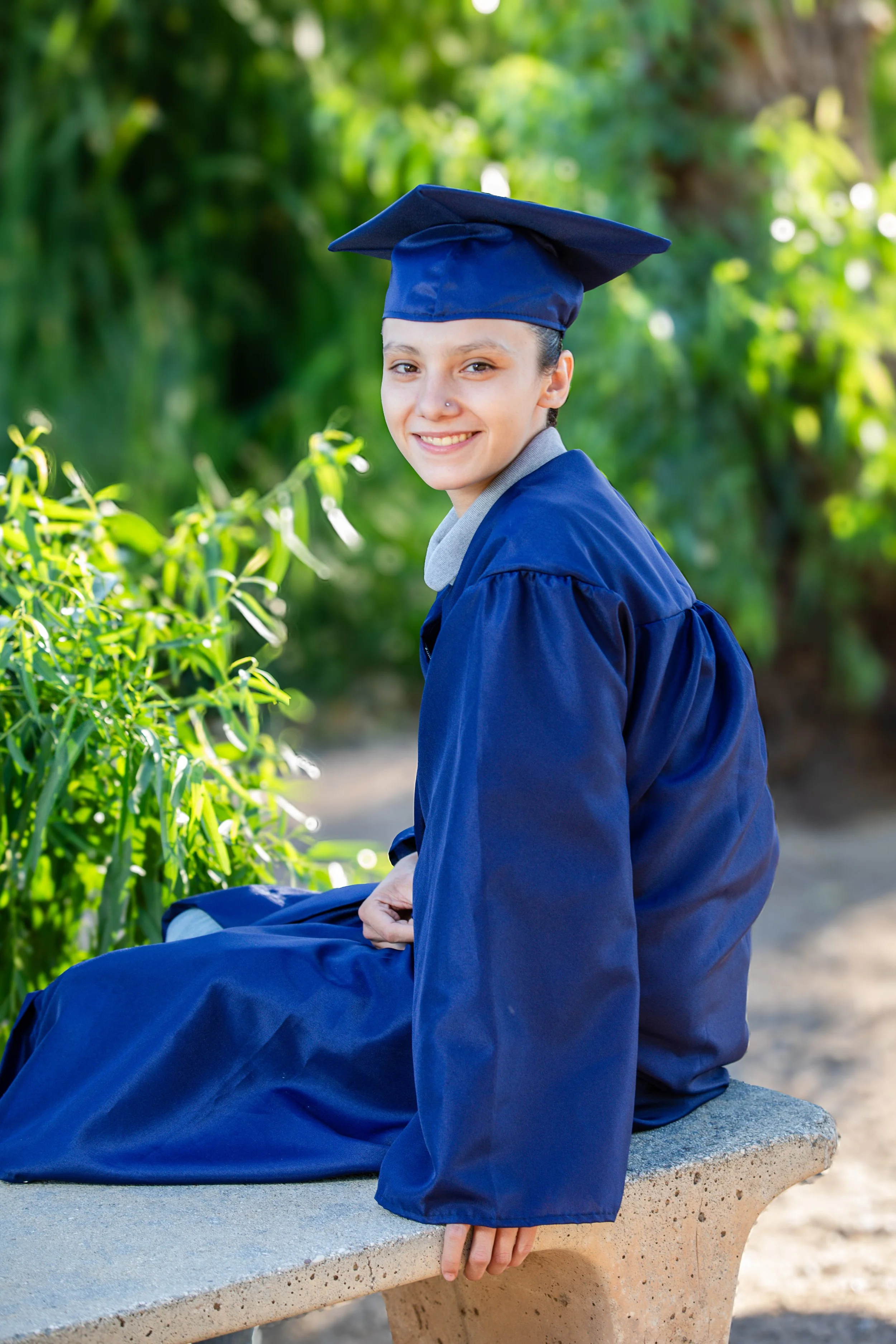 Young woman in a blue graduation gown and cap sitting on a concrete bench outdoors, smiling, with greenery in the background.