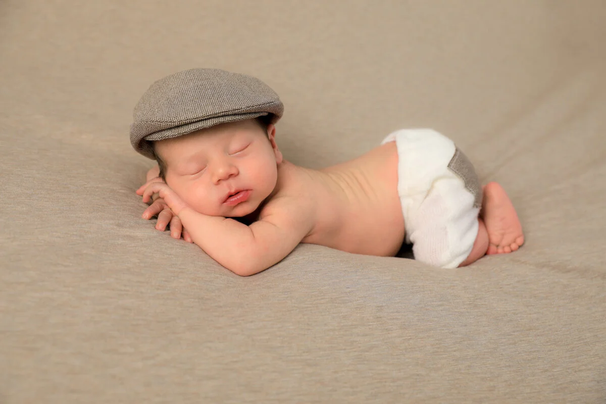 Newborn baby wearing a flat cap and diaper, sleeping on its stomach on a beige surface.