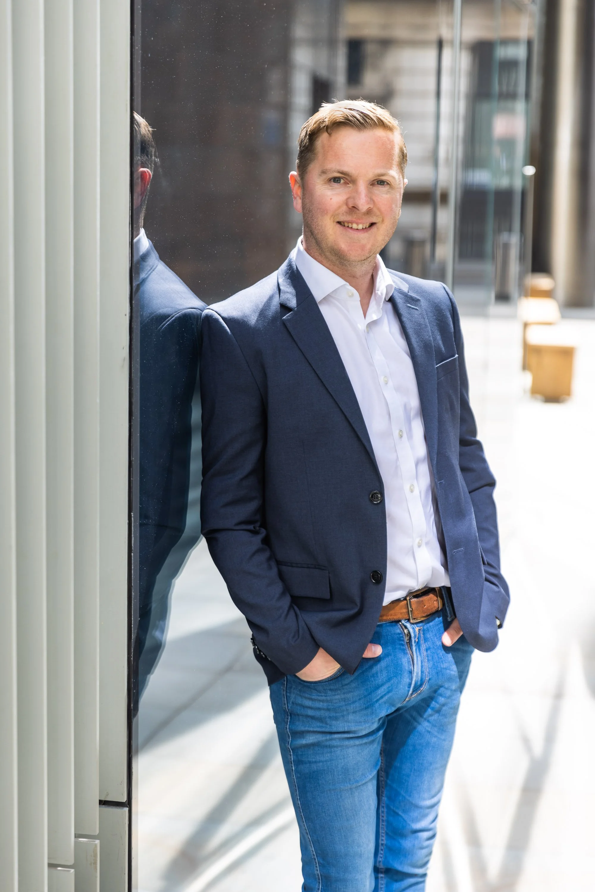 Corporate photography showing a man in business attire standing outdoors, smiling, with hands in pockets, leaning against a glass wall.