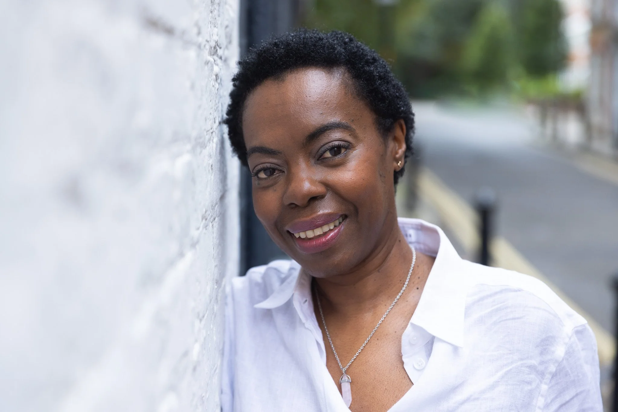 A smiling person wearing a white shirt and necklace leans against a white brick wall outdoors.