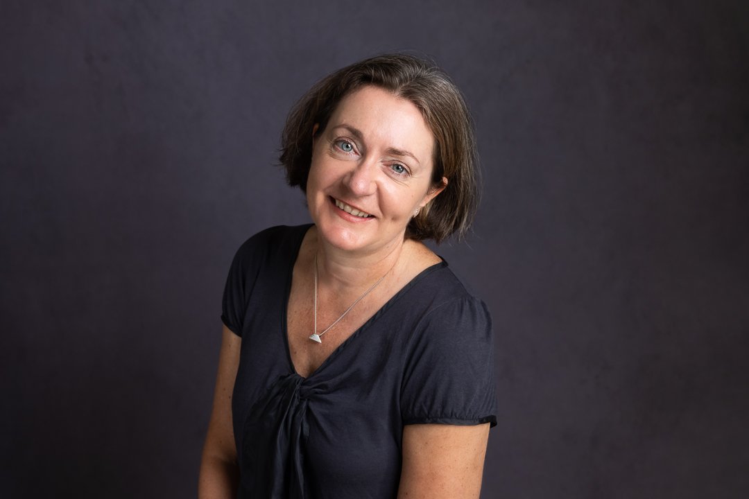 Smiling woman with short dark hair wearing a black blouse and necklace, against a dark background.