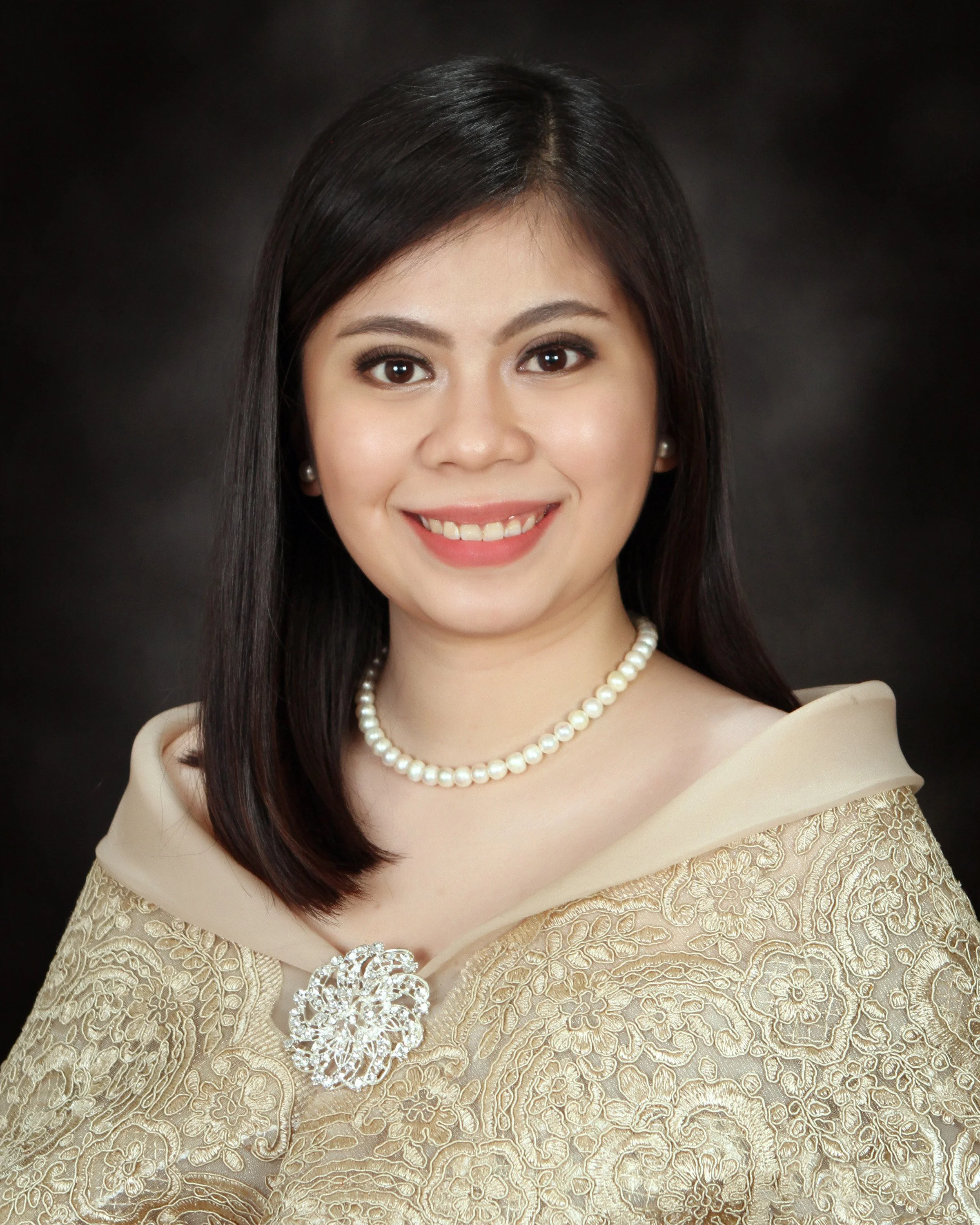 A woman with straight dark hair, wearing pearl earrings and necklace, dressed in an ornate gold traditional outfit with a brooch, smiling at the camera against a dark background.