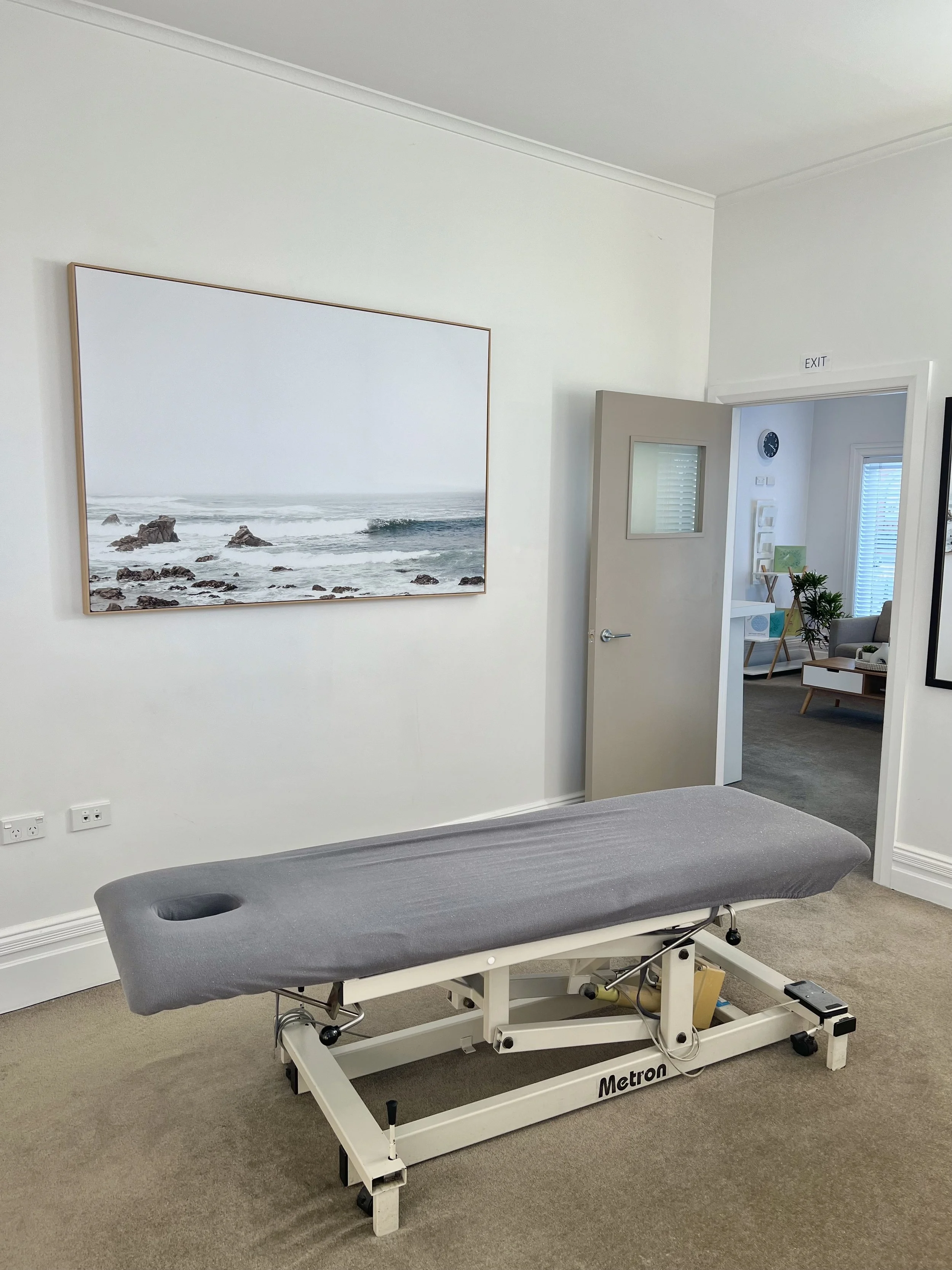 A therapy or massage table with a gray cover in a room with white walls and beige carpet, a large framed photo of the ocean on the wall, an open door leading to a waiting area with a clock, plants, and seating.