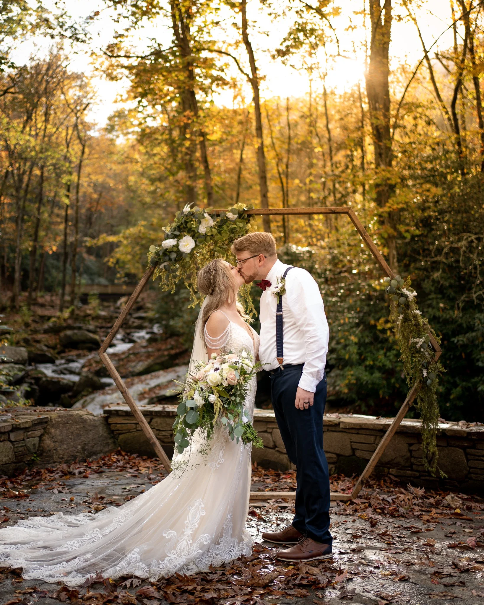 A bride and groom share a kiss outdoors in autumn, standing under a wooden floral arch, surrounded by fallen leaves and trees with golden leaves.