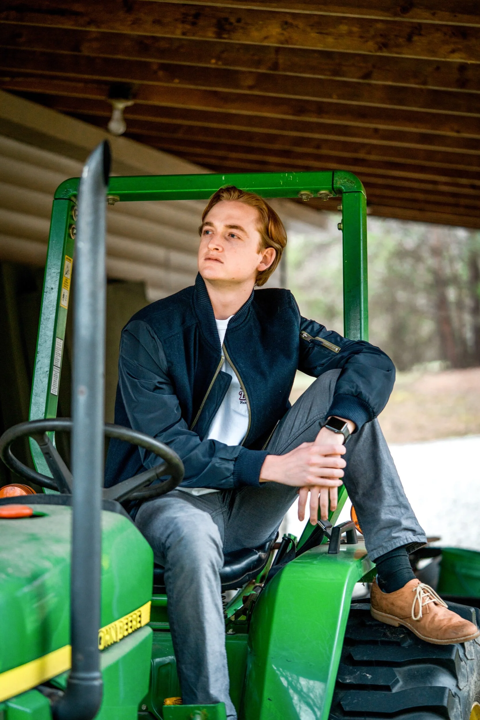 Young man sitting on a green John Deere tractor, wearing a dark blue jacket, gray pants, and brown boots, looking thoughtfully to the side under a wooden roof.
