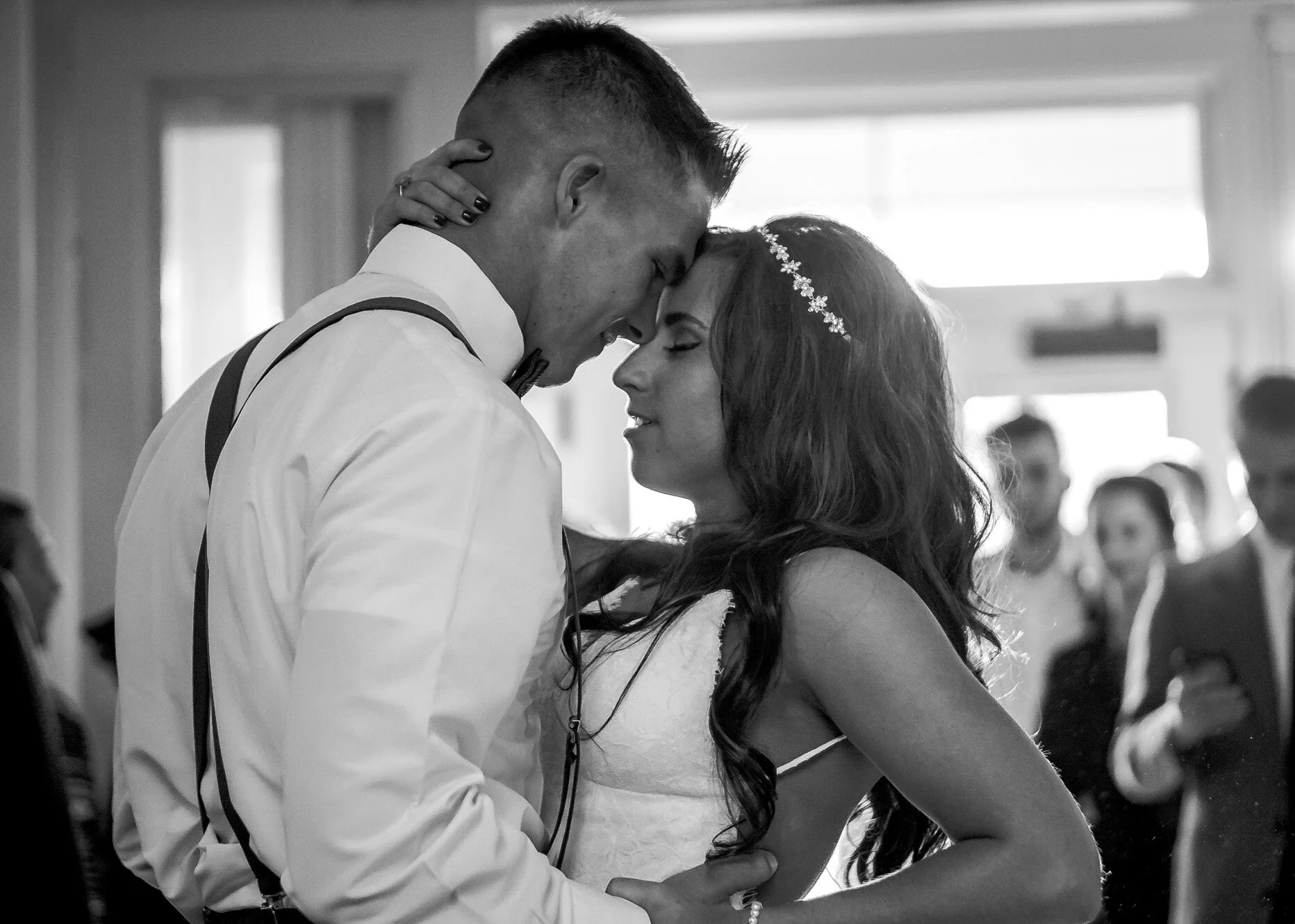 A black and white photograph of a newlywed couple sharing a dance at their wedding reception, with guests in the background.