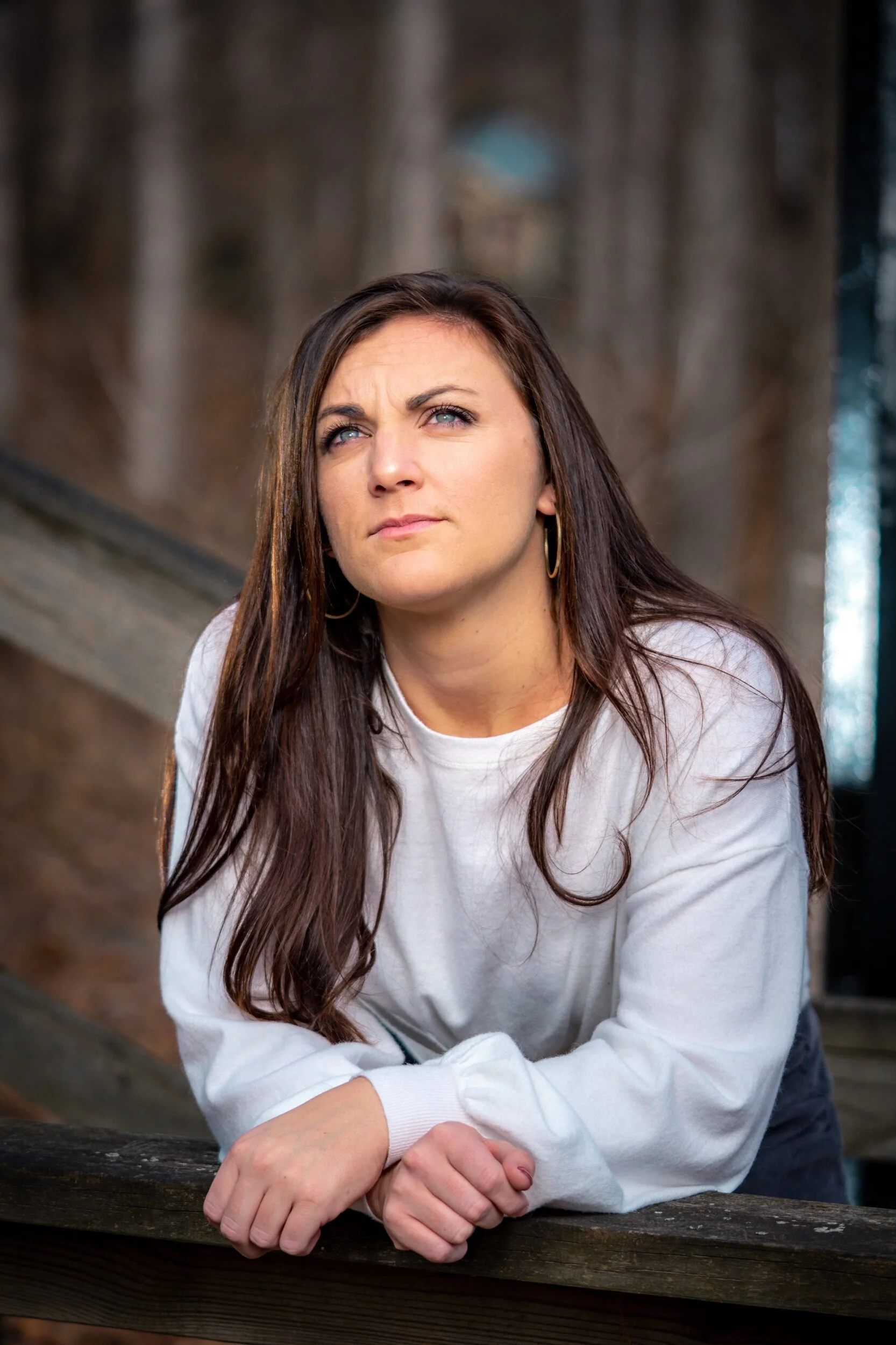 A woman with long brown hair, wearing a white sweatshirt and gold hoop earrings, leaning on a wooden railing outdoors, looking contemplative with a serious expression, background of blurred trees.