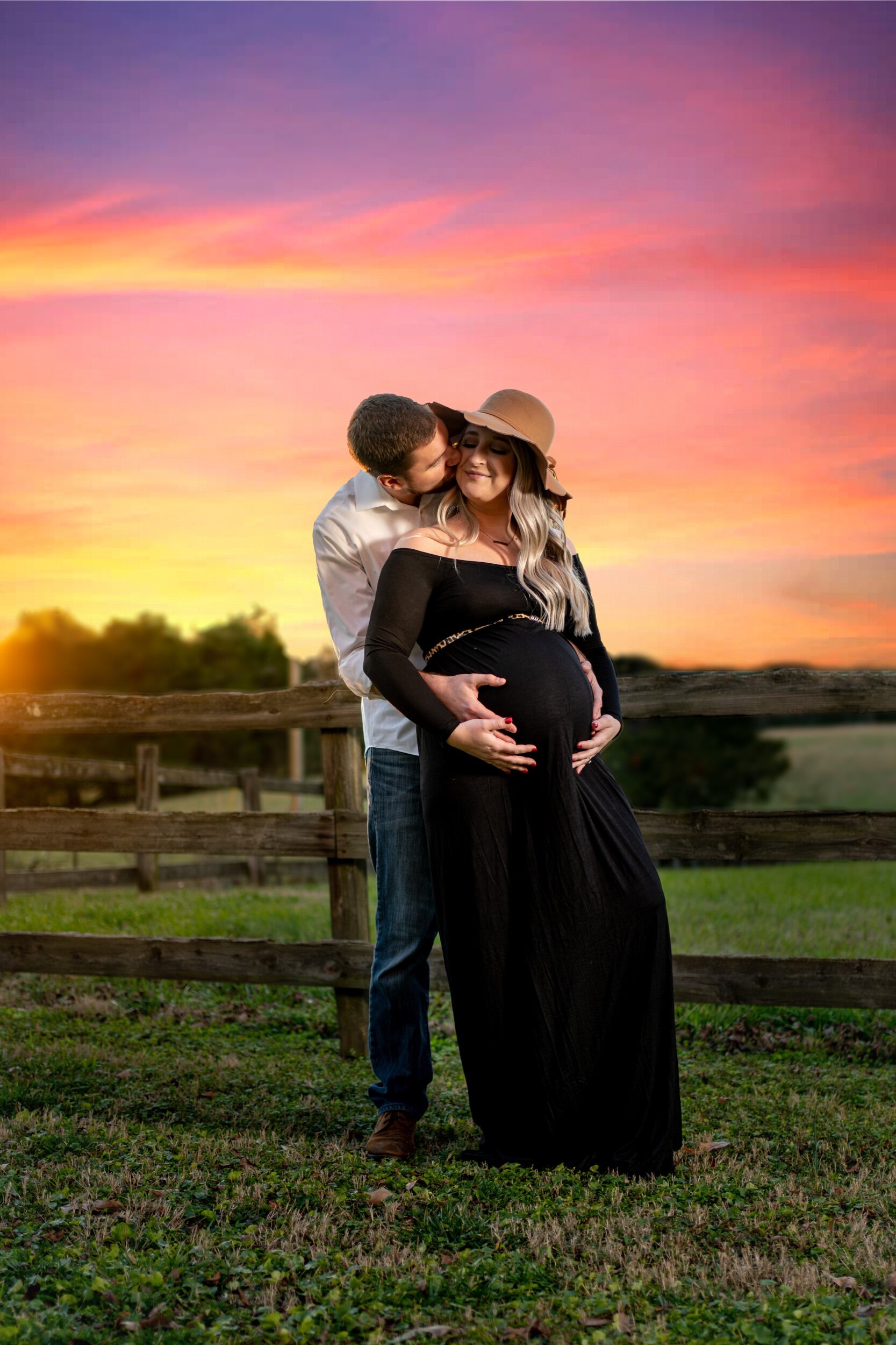 A pregnant woman with long blonde hair wearing a black dress and a beige sun hat, standing outdoors on grass near a wooden fence at sunset, with her partner, a man in a white shirt and jeans, embracing her from behind and kissing her cheek.