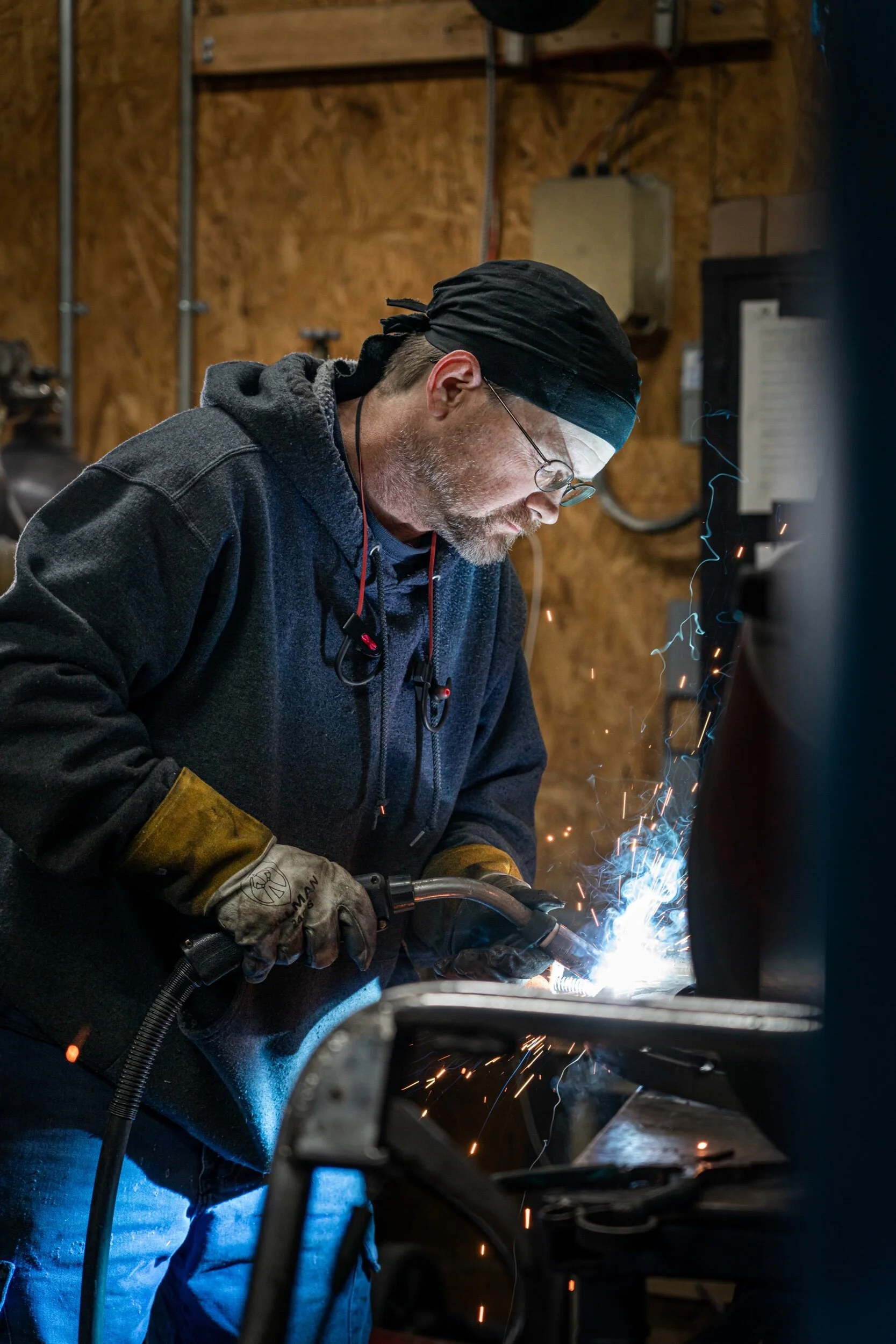 A man welding metal inside a workshop, wearing a black bandana, glasses, and protective gloves.