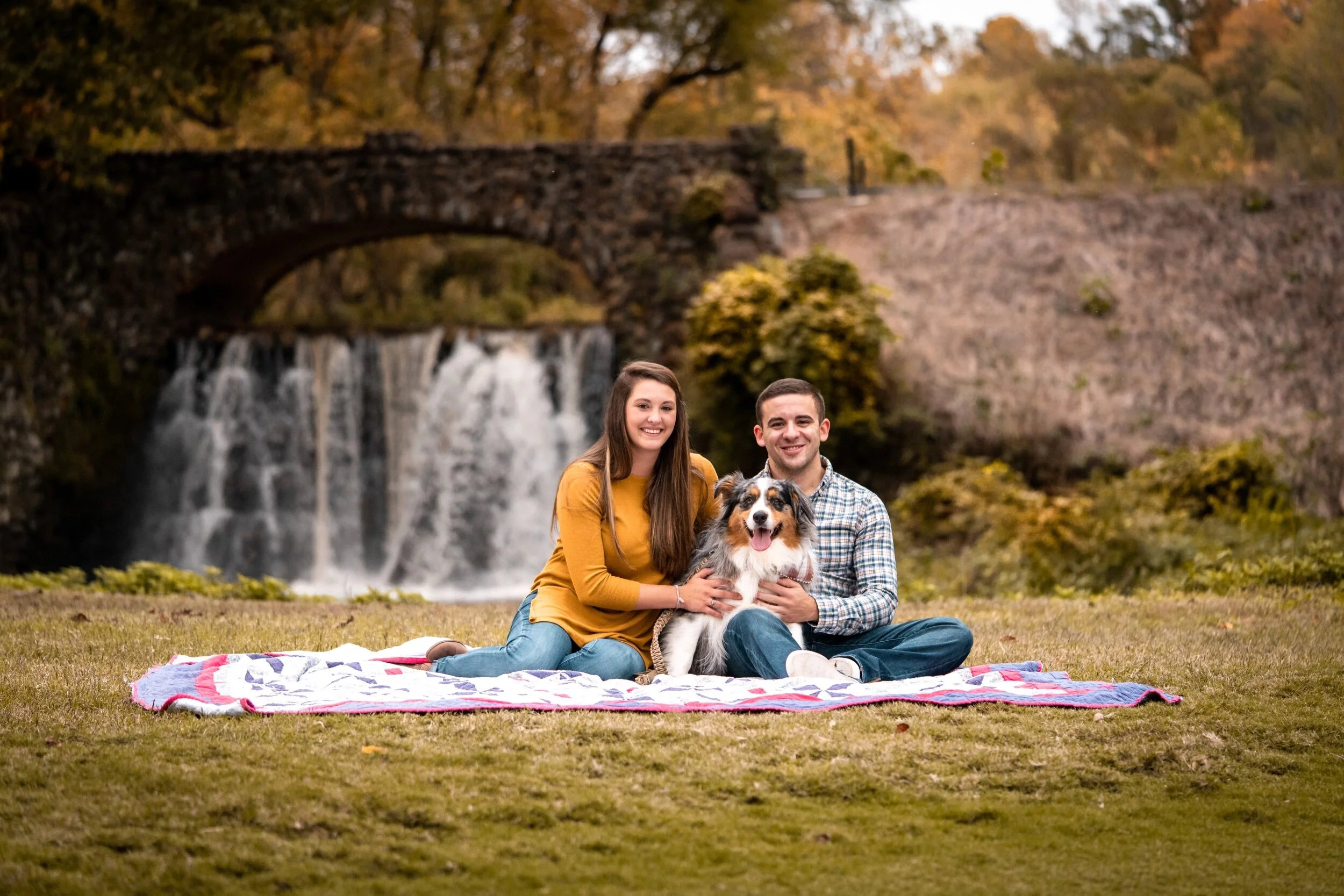 A smiling couple and their dog sitting on a blanket outdoors with a waterfall and trees in the background during autumn.