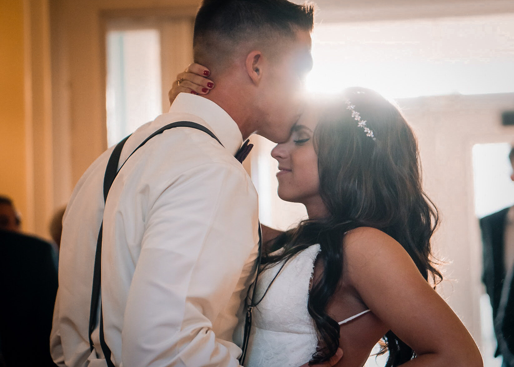 A couple, dressed in wedding attire, sharing a dance at their wedding reception, with bright sunlight shining behind them.