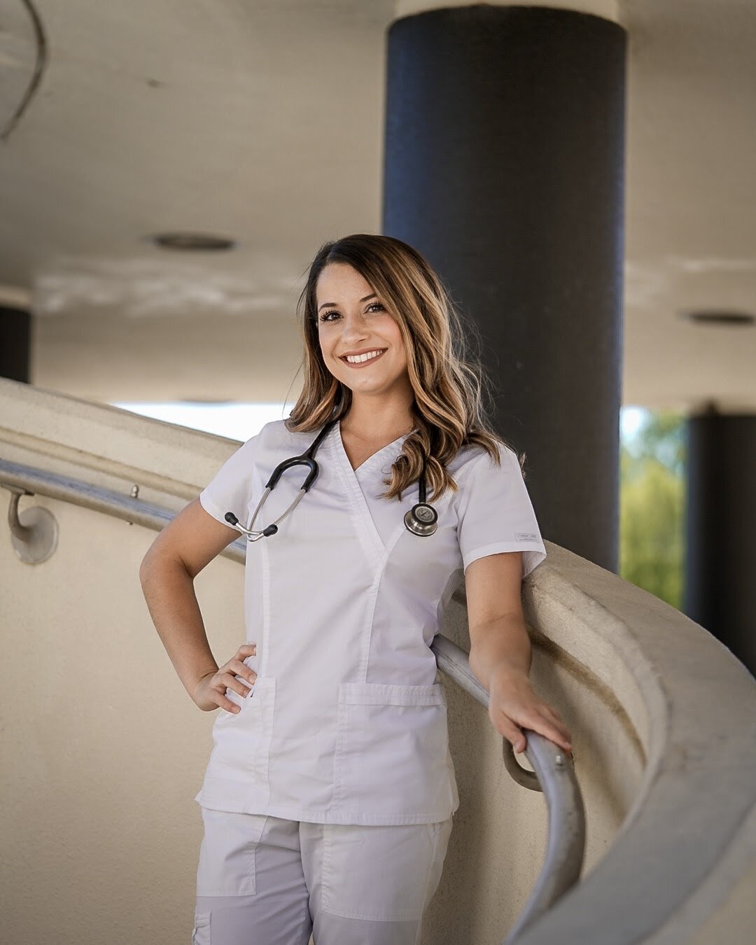 A female healthcare professional smiling, wearing white scrubs and a stethoscope, standing outdoors by a curved railing with large black columns in the background.