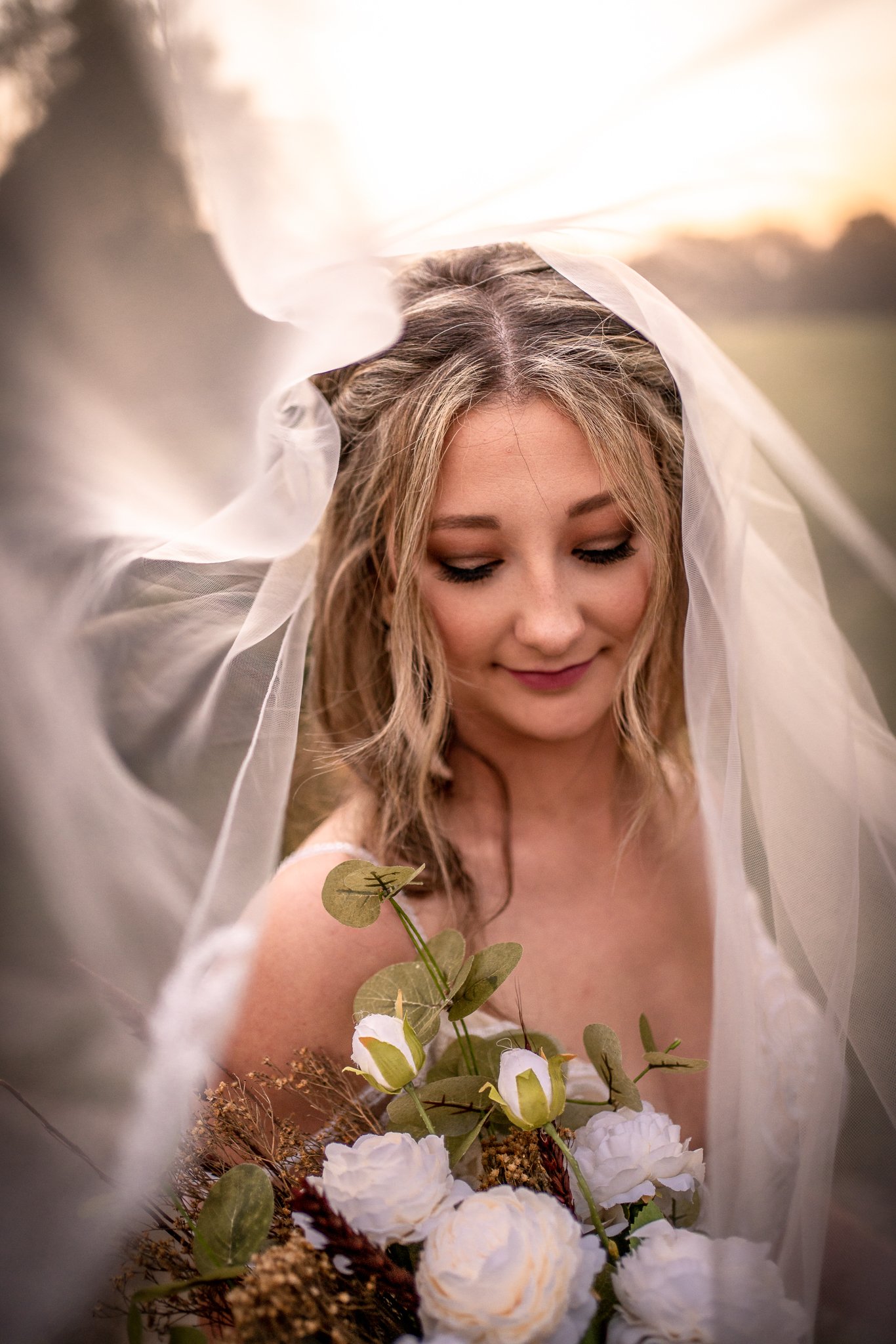 A bride with a veil and makeup, holding a bouquet of white roses and greenery, outdoors at sunset.