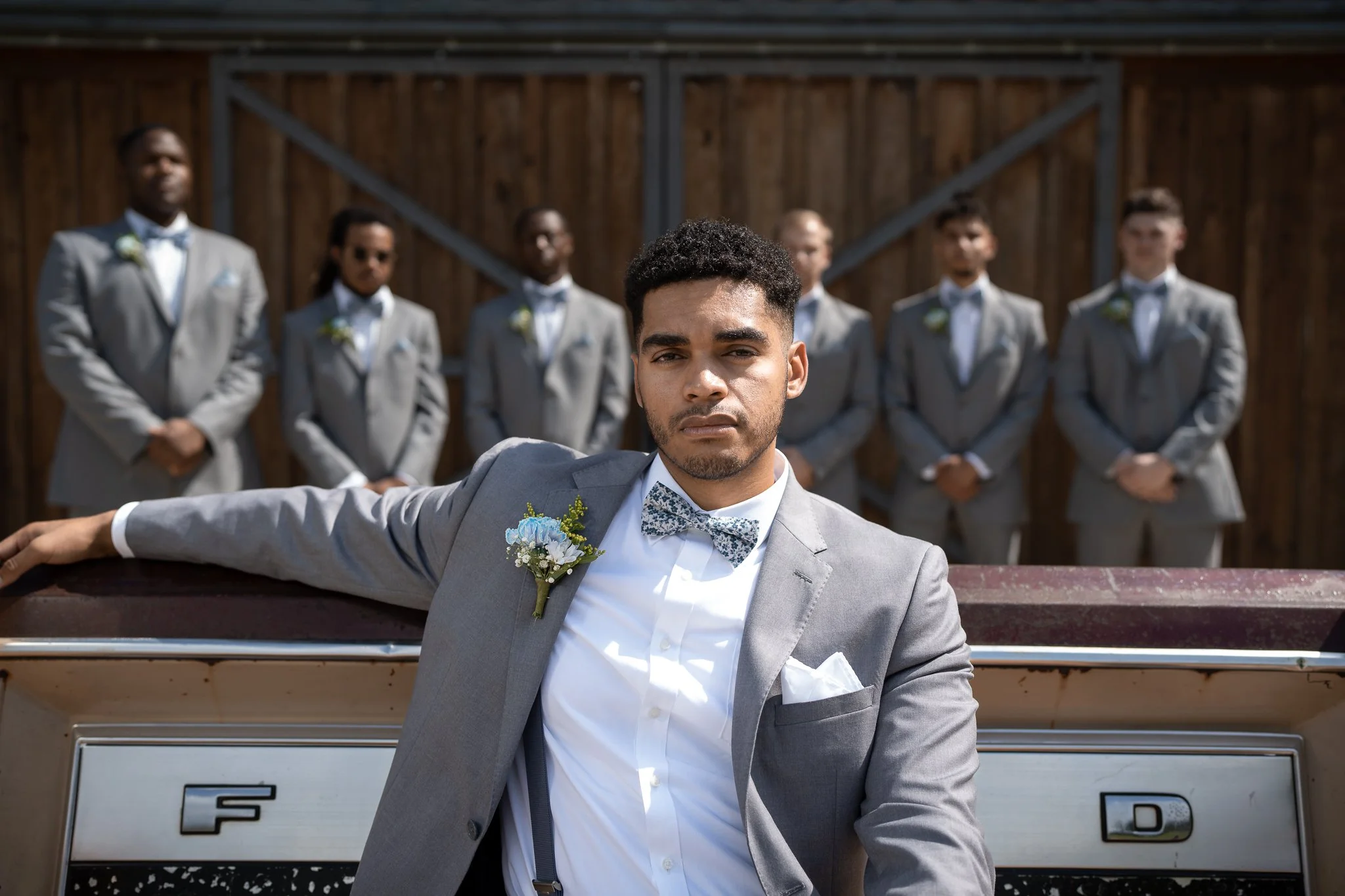 A man in a gray suit with a bowtie and boutonniere sitting on a vehicle, with seven men in matching gray suits and bowties standing behind him outdoors in front of a wooden fence.
