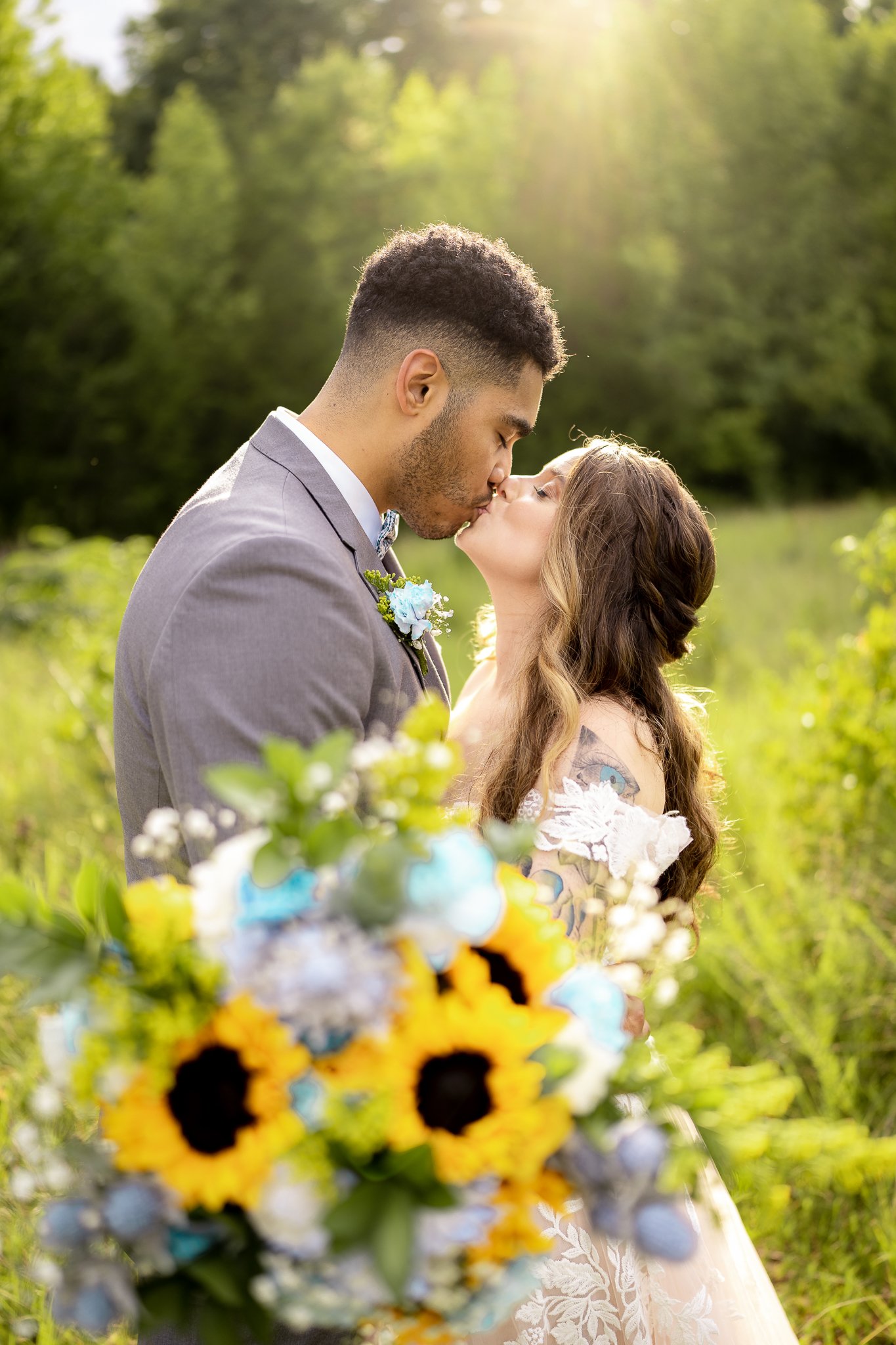 A couple kissing outdoors with a sunlit green forest background, a vibrant bouquet of sunflowers and blue flowers in the foreground.