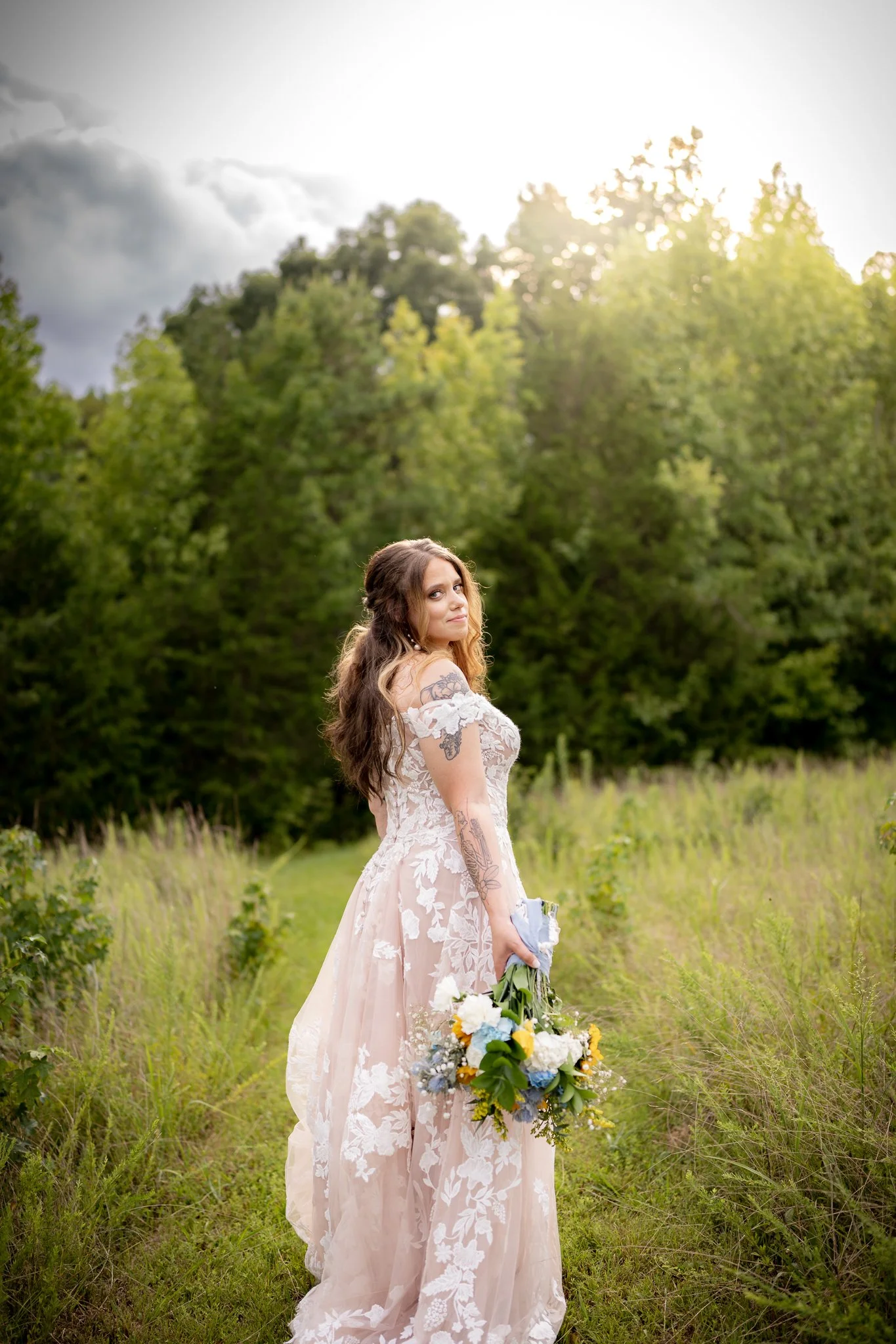 A woman in a white lace wedding dress holding a bouquet of flowers standing in a grassy field with trees in the background during sunset.