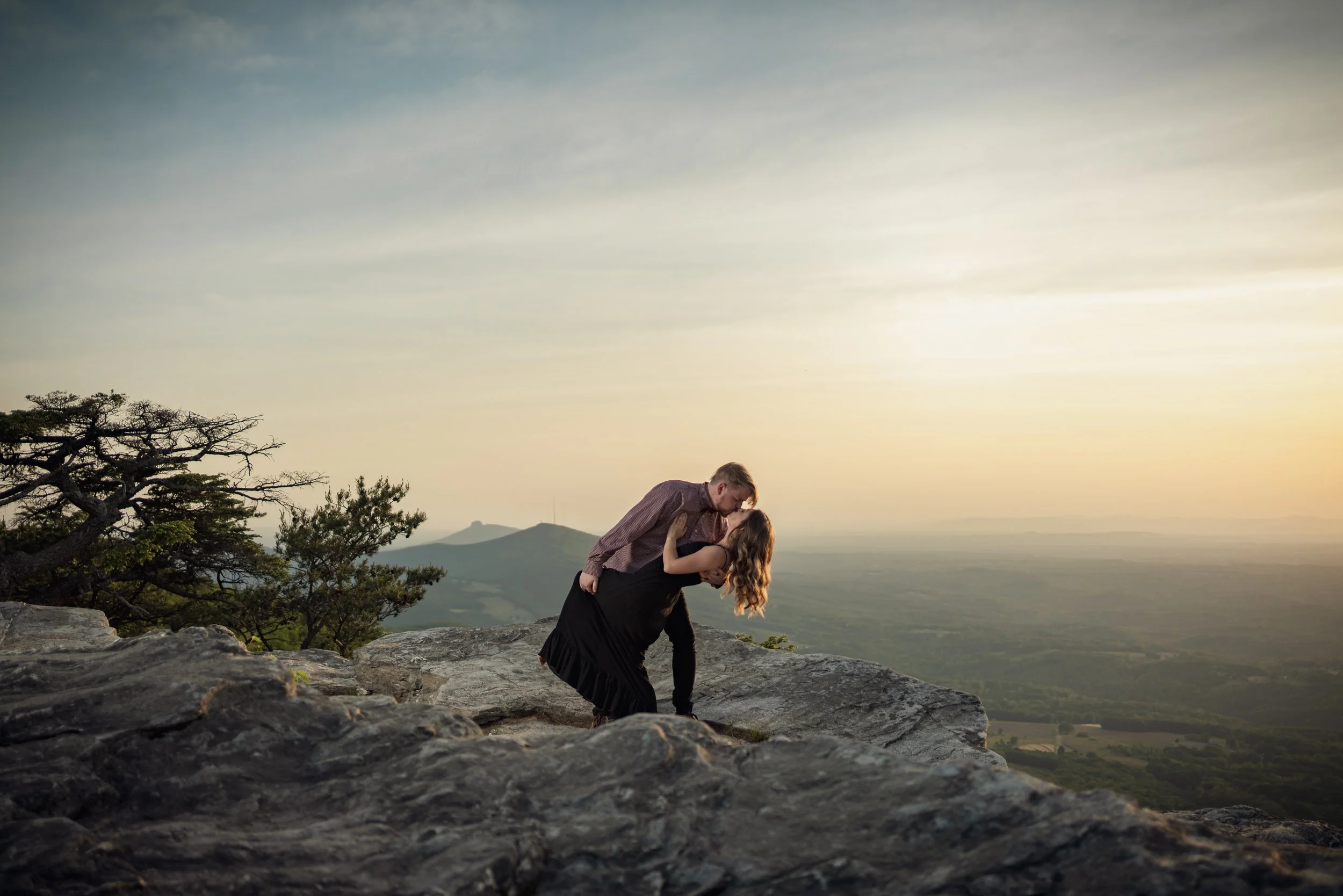 A couple sharing a kiss on a rocky mountaintop during sunset with a landscape view in the background.
