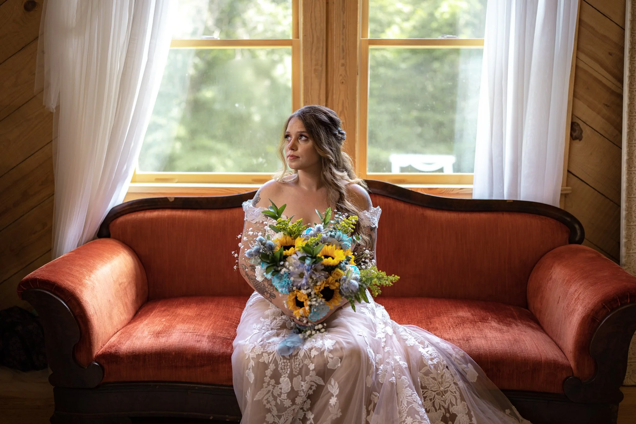 A bride with long wavy hair, wearing an off-shoulder lace wedding gown, sitting on an orange vintage sofa, holding a bouquet of yellow sunflowers and blue flowers, inside a wooden room with large windows and white curtains.