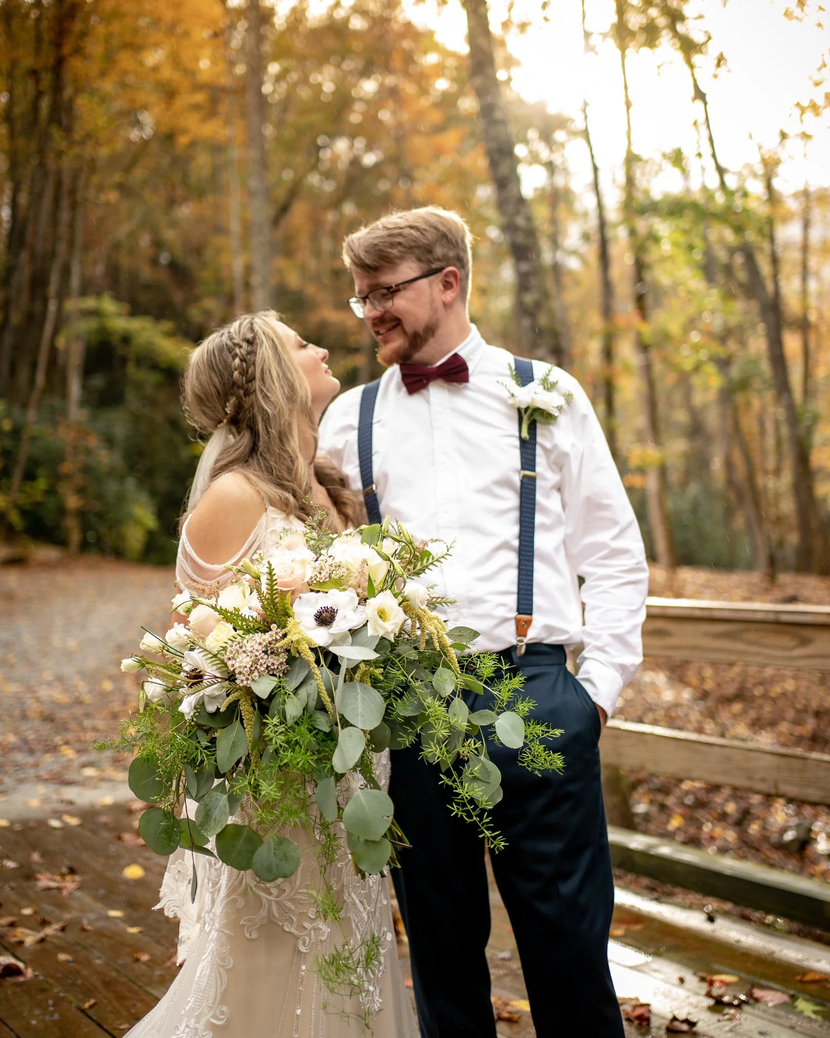 A bride and groom stand close and look at each other smiling in an autumn forest setting. The bride holds a large bouquet of white and light pink flowers with greenery. The groom wears a white shirt with suspenders, a bow tie, and a boutonniere. Sunl