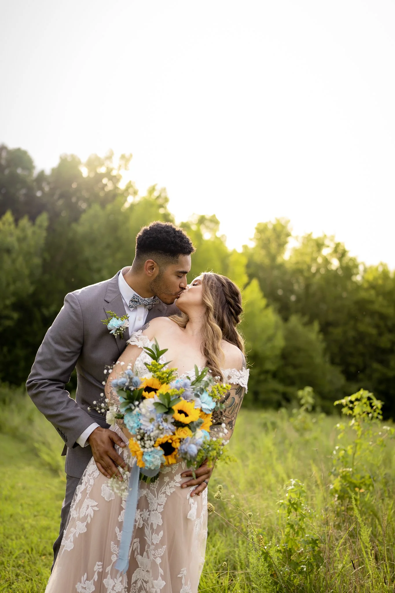 A newlywed couple sharing a kiss outdoors, the groom in a gray suit with a bowtie, and the bride in a lace off-the-shoulder wedding dress holding a bouquet of yellow, blue, and white flowers, in a green field with trees in the background, during suns
