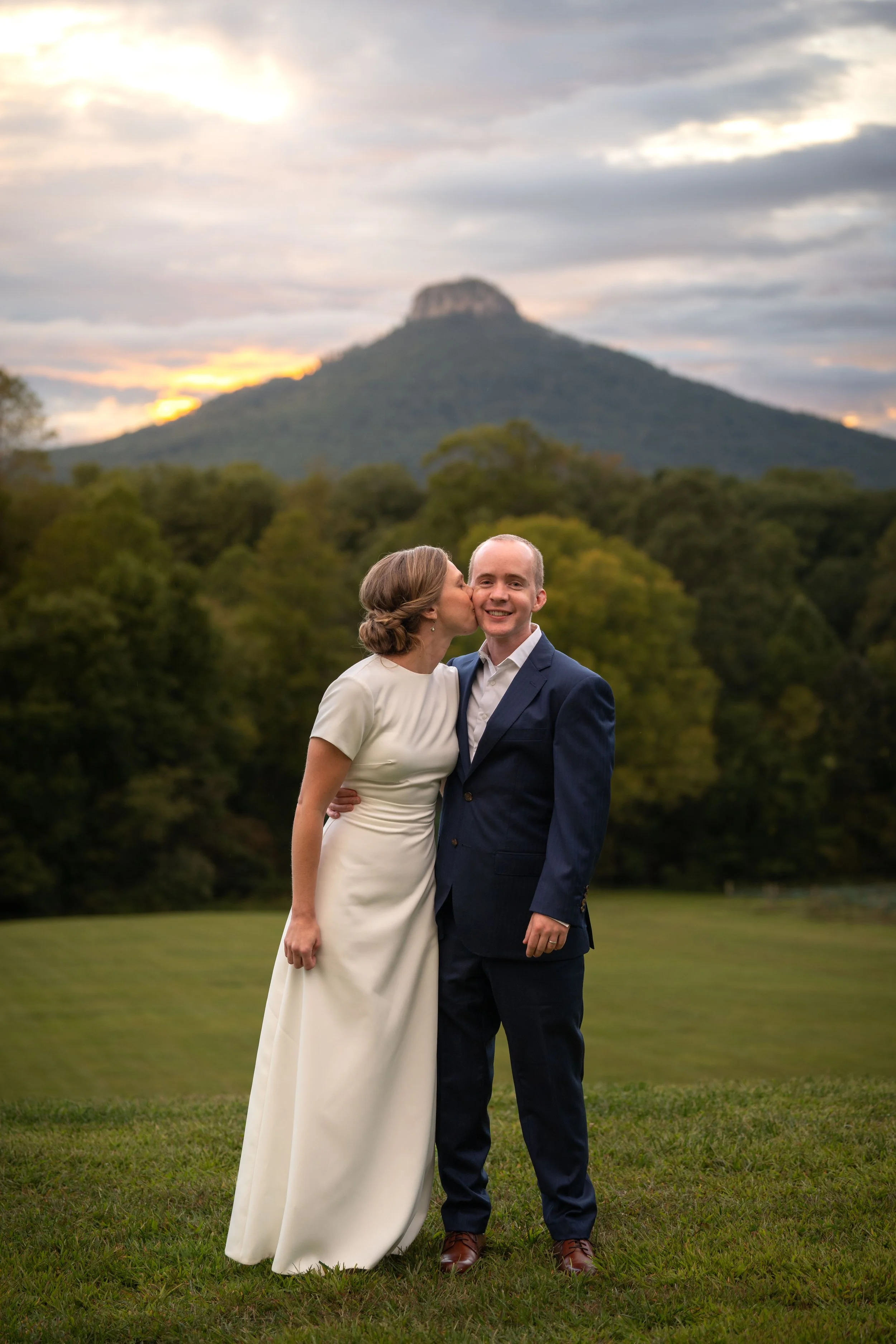 A woman in a white dress kisses a man in a suit on the cheek outdoors, with a mountain and cloudy sky in the background during sunset.