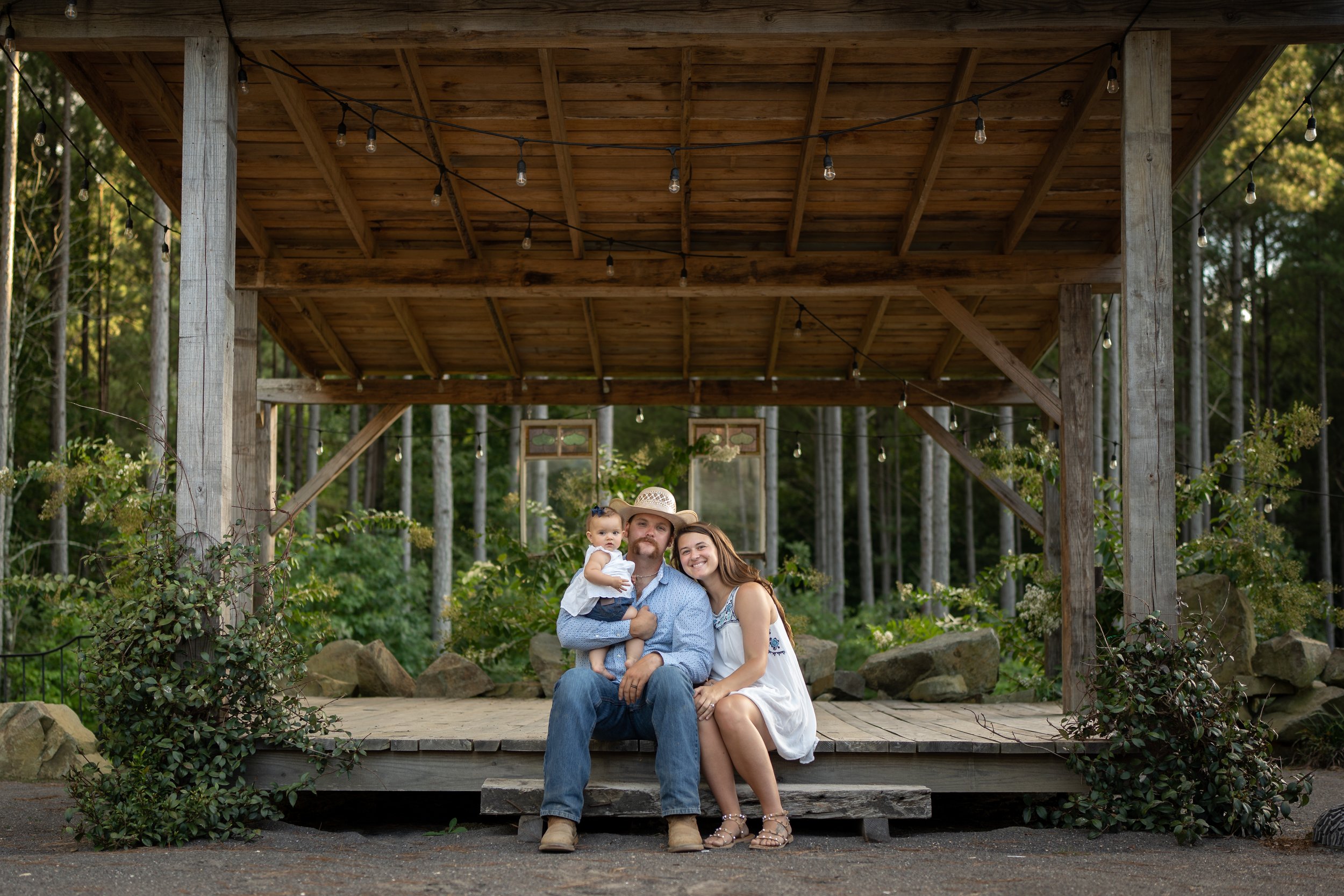 A family of three sitting on a small wooden stage outdoors, surrounded by trees and rocks, with string lights hanging from a wooden pavilion ceiling.