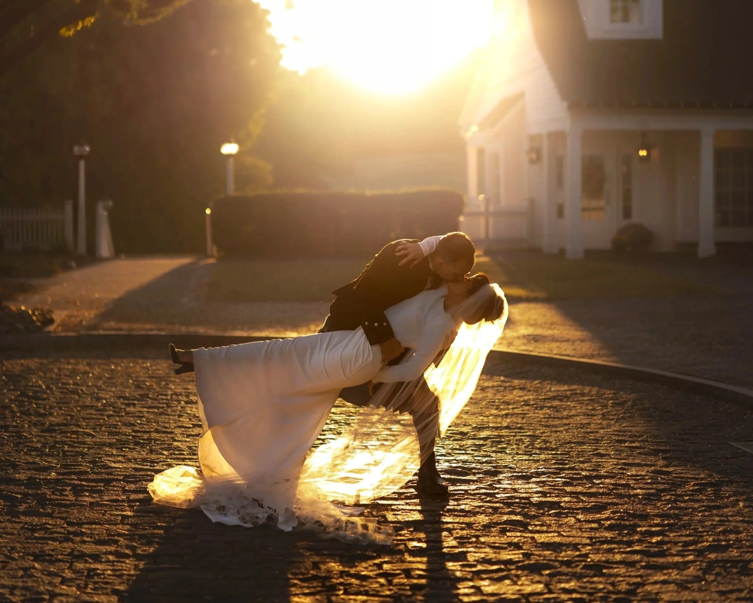 A couple, dressed in wedding attire, sharing a kiss on the ground during sunset, with a house and street lamps in the background.