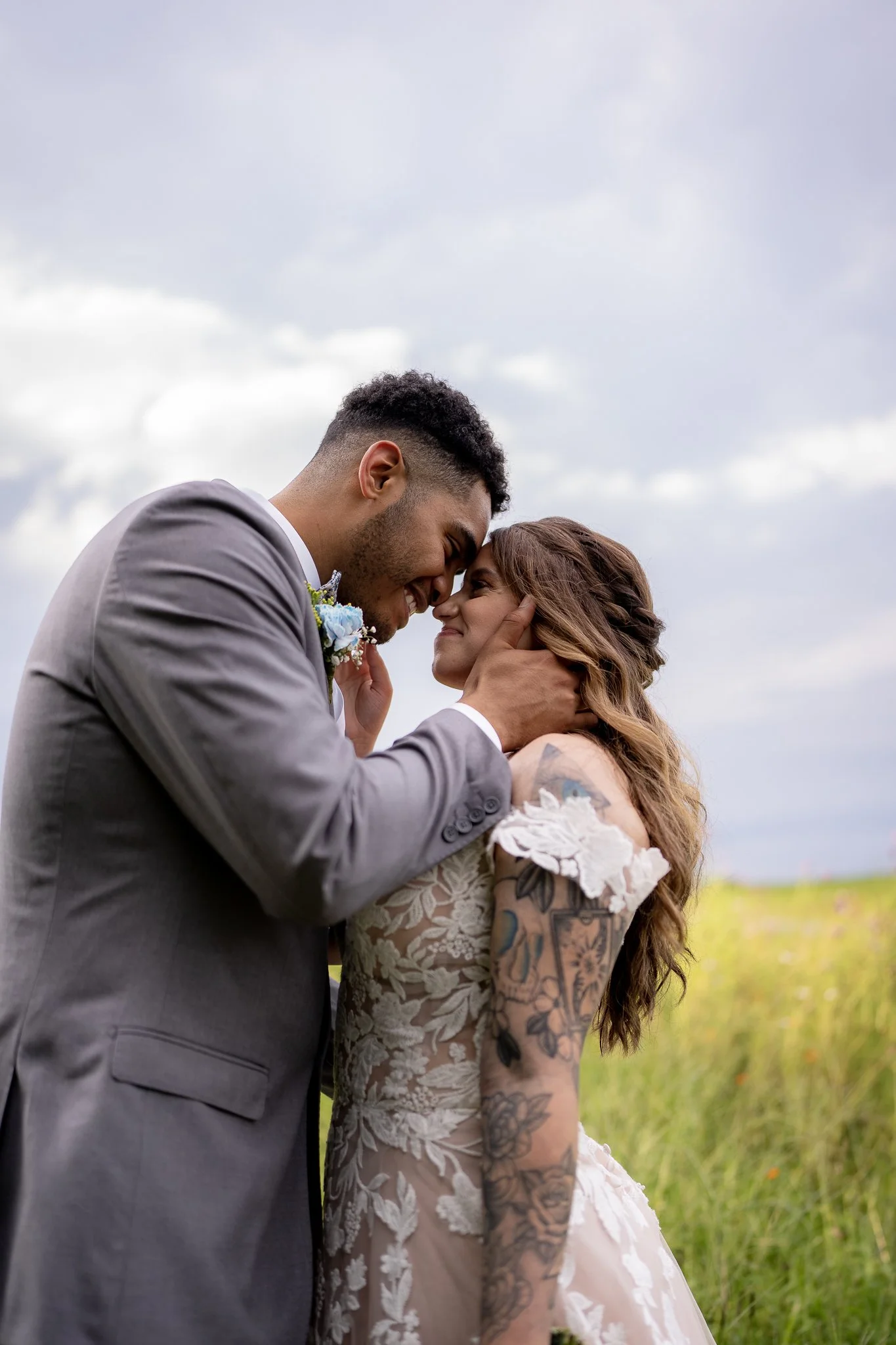 A bride and groom sharing a romantic moment outdoors, touching foreheads, with a cloudy sky and grassy field in the background.